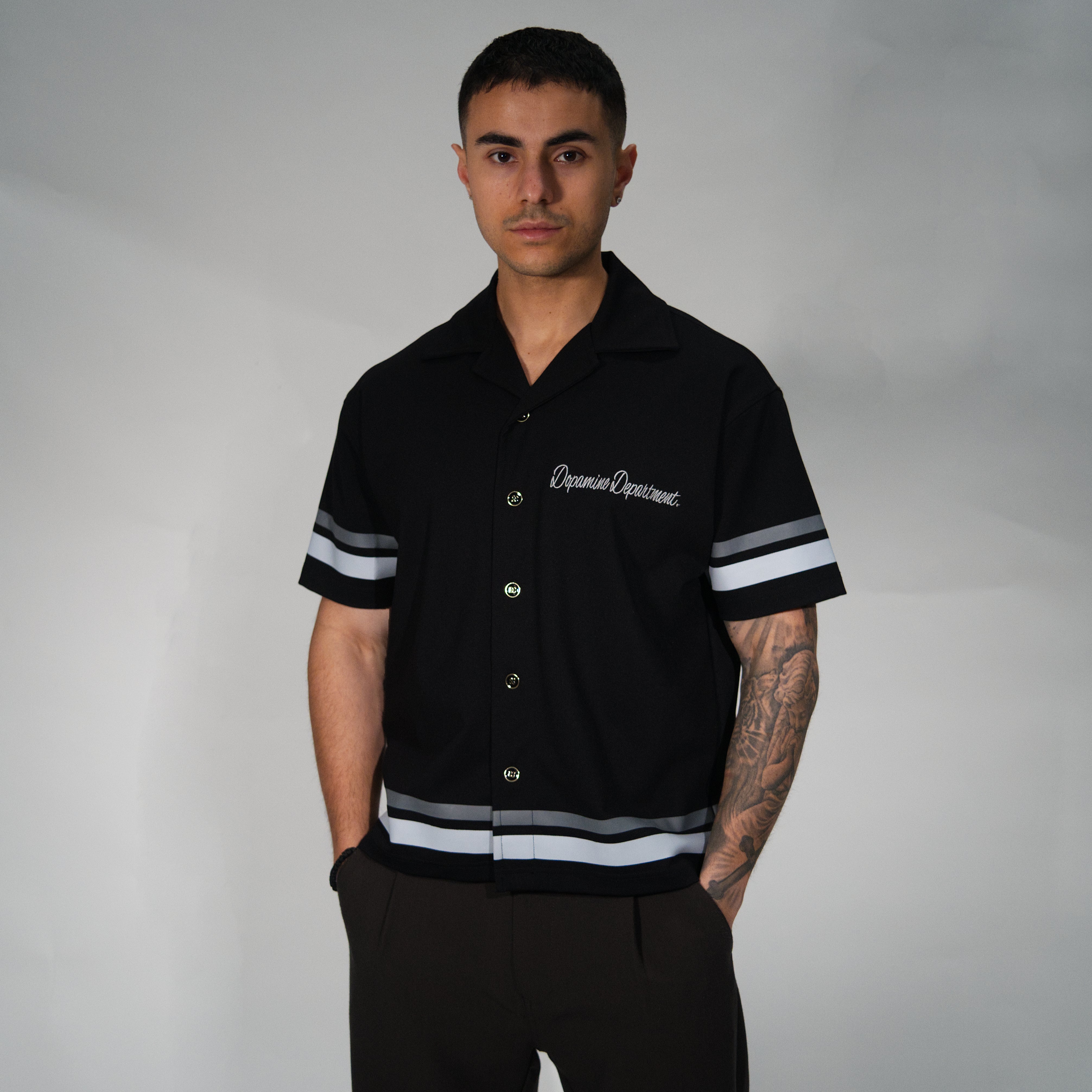 A man wearing a black short-sleeve button-up shirt with white and grey striped ribbed cuffs and hem, featuring 'Dopamine Department' in white script on the chest, posed against a neutral grey studio background.