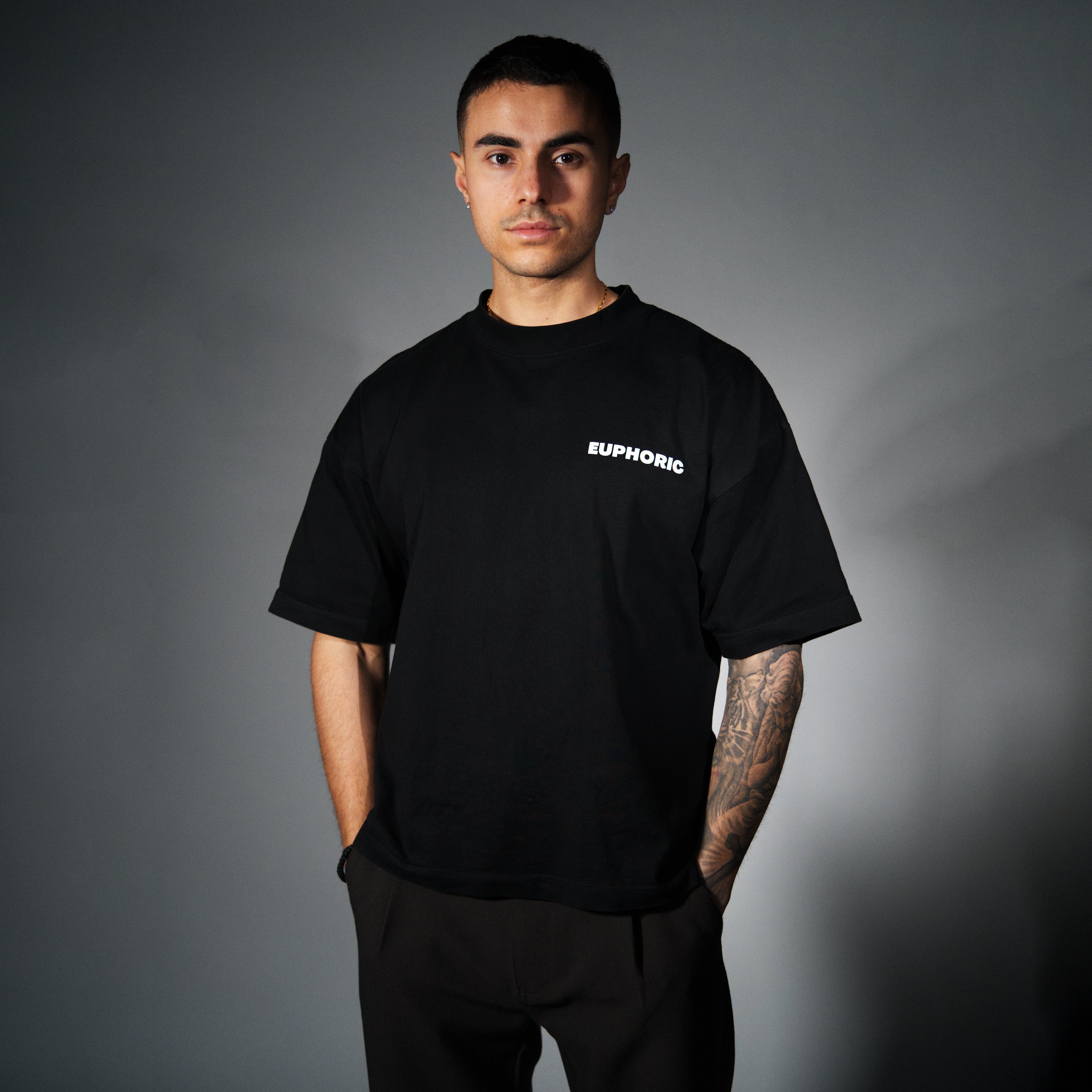 A man wearing a black short-sleeve t-shirt with the brand name 'EUPHORIC' printed in white on the left chest, paired with black pants, set against a dark grey studio background.