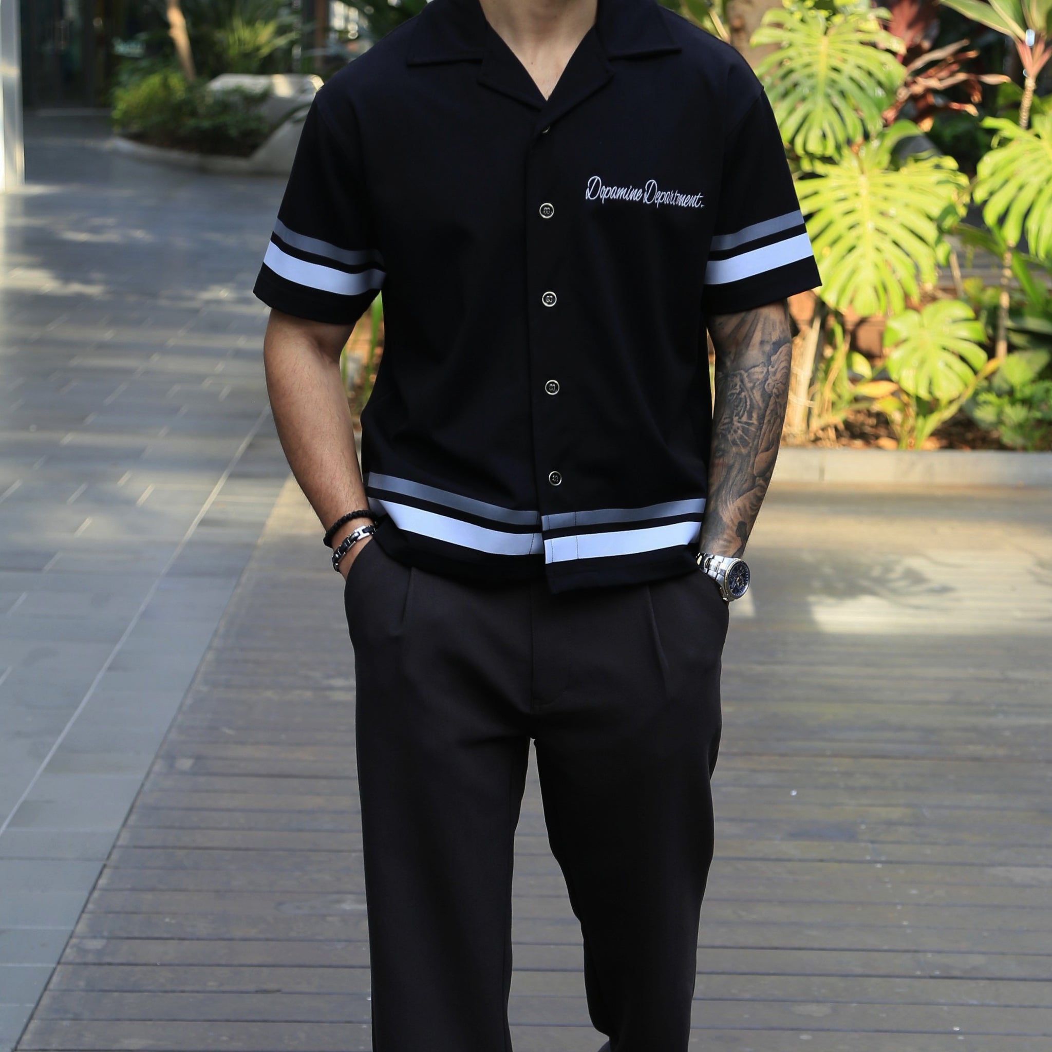 Person wearing a Euphoric Black Relaxed Button Shirt standing outdoors on a path with greenery.