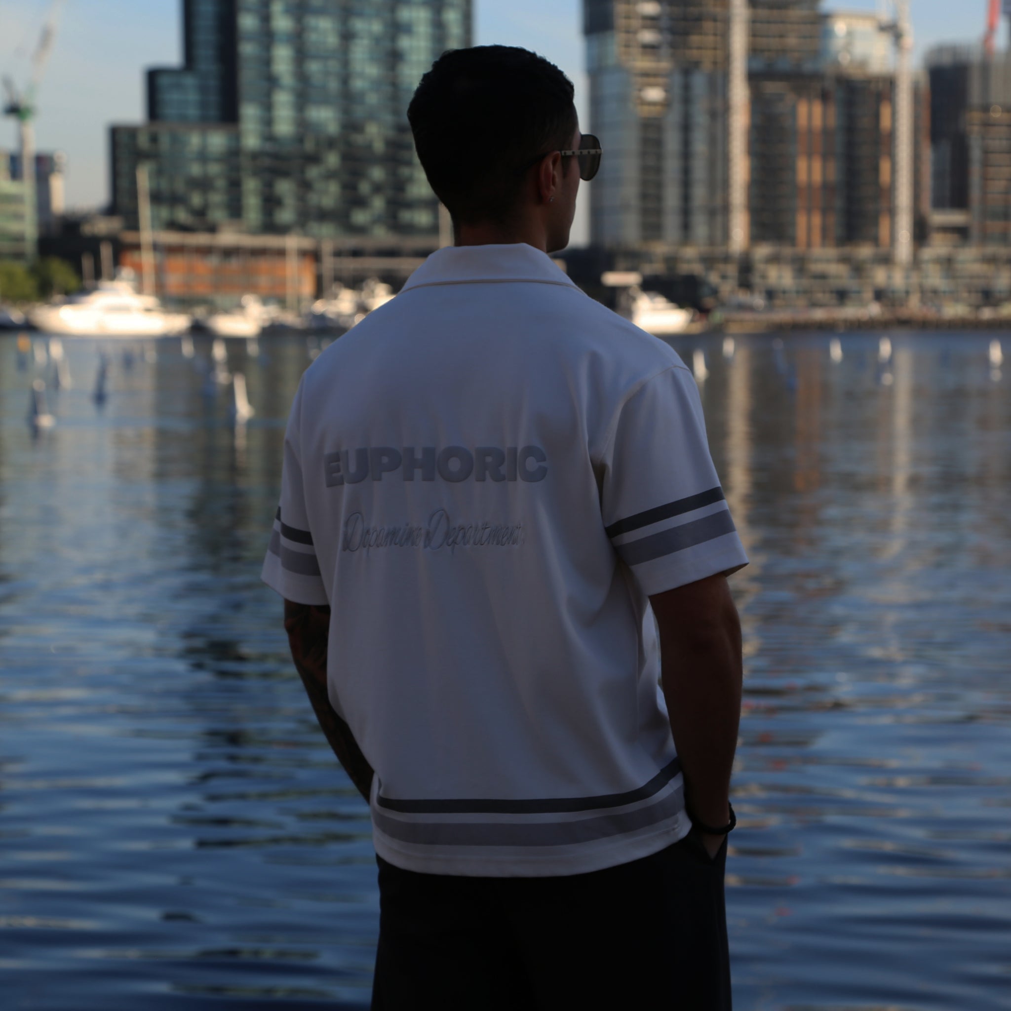 Man wearing a euphoric relaxed white button shirt standing by a waterfront with city skyline in the background.