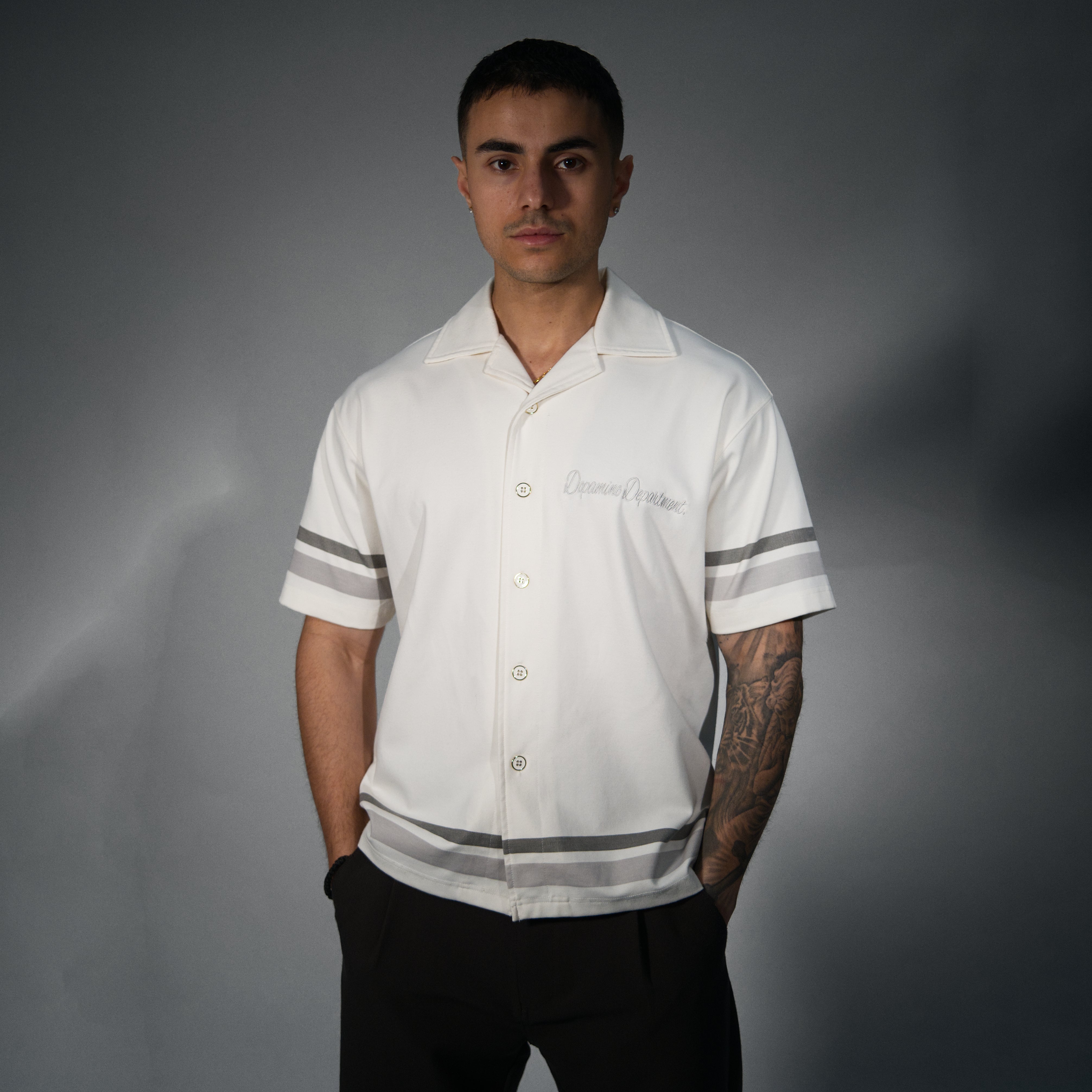 A man wearing a white short-sleeve button-up shirt with grey stripes on the sleeves and hem, paired with black pants, standing against a dark grey studio background.
