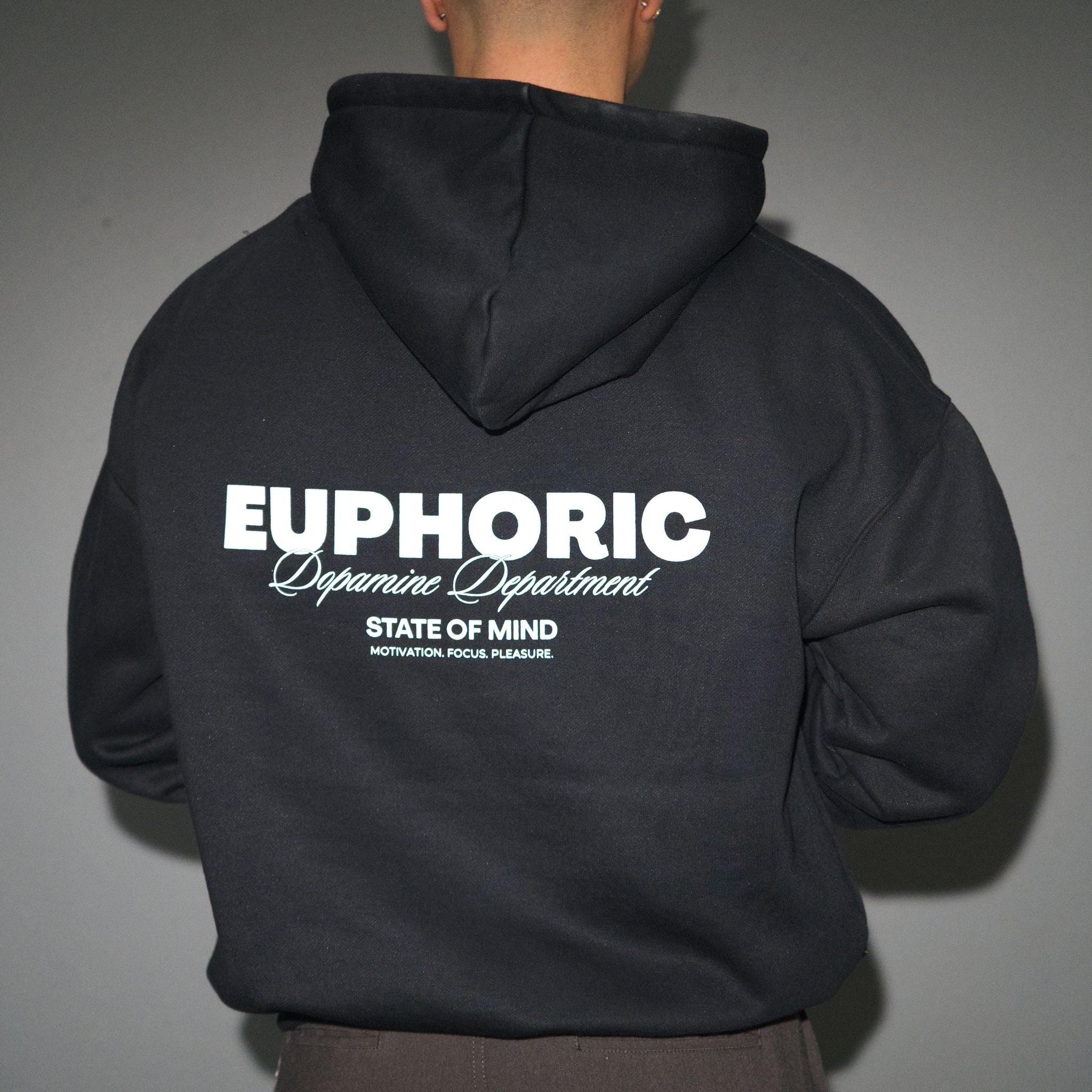A person shown from the back wearing a black hoodie with bold white text on the back reading 'EUPHORIC' and 'Dopamine Department' plus smaller text below, against a plain grey wall.
