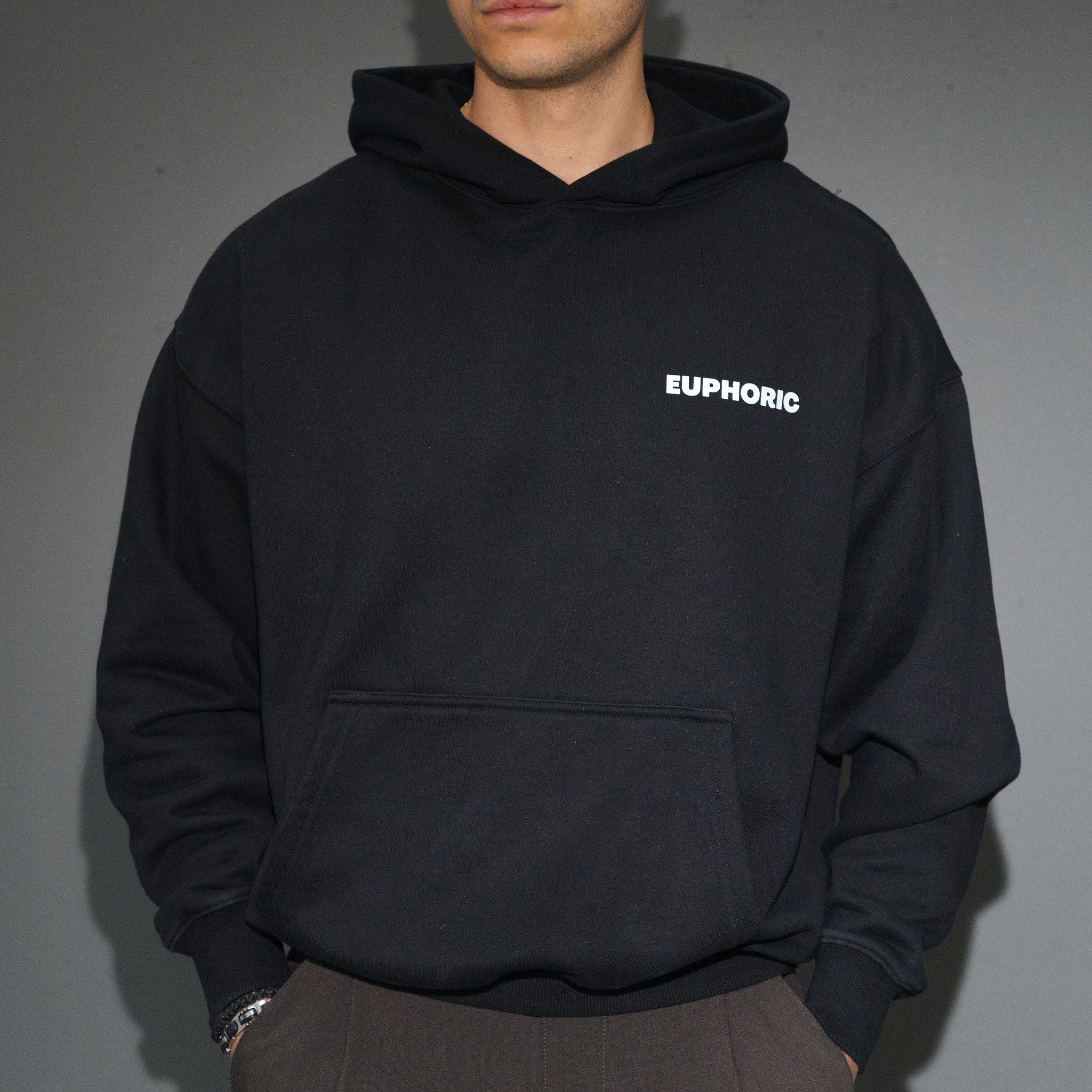 A person wearing a black pullover hoodie with the word 'EUPHORIC' printed in white on the left chest, paired with brown pants against a plain grey background.