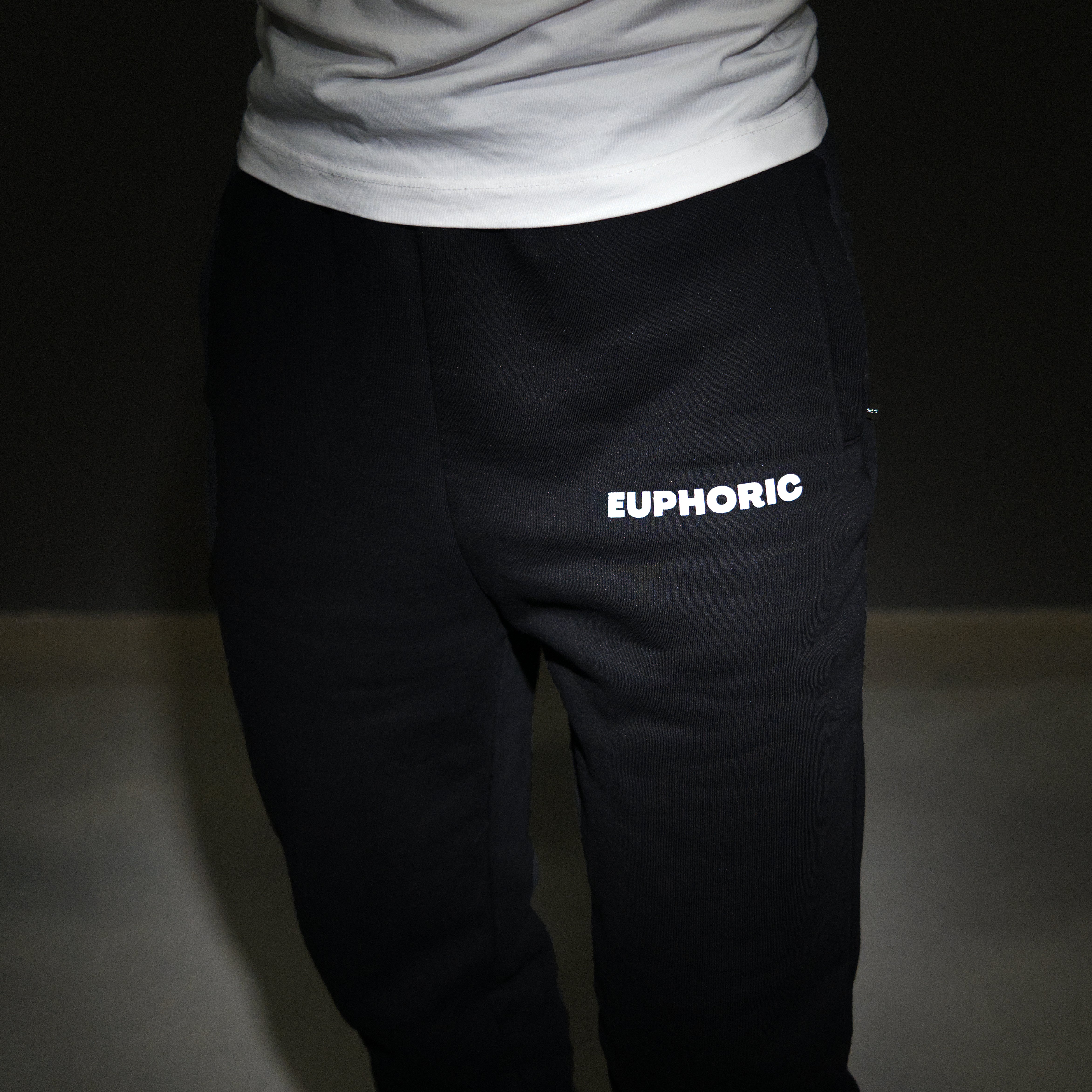 Black sweatpants worn by a model, featuring the word 'EUPHORIC' printed in white capital letters on the left thigh, paired with a white t-shirt against a dark studio background.