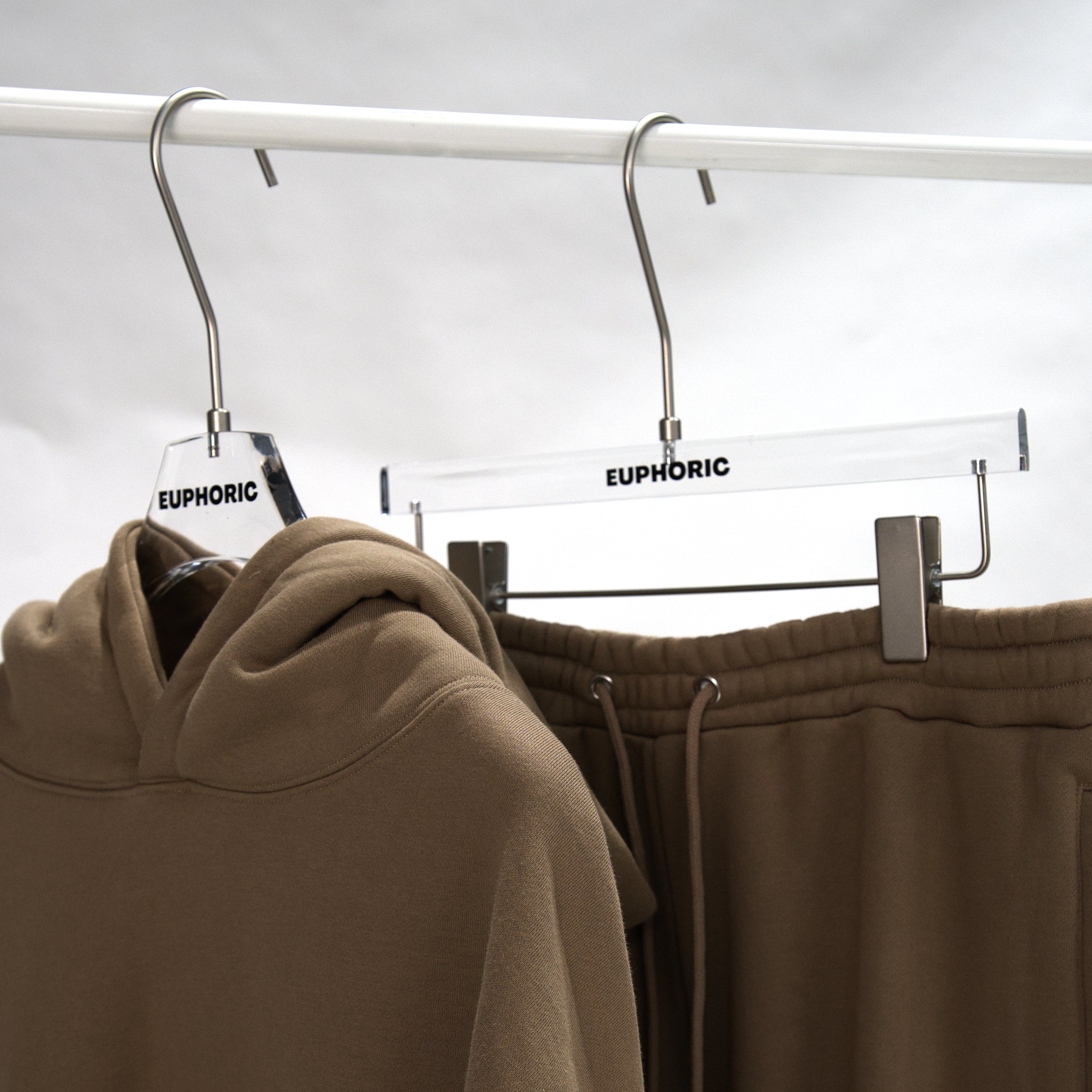 A taupe-brown hoodie and matching sweatpants hang on clear acrylic hangers with the brand name 'EUPHORIC' printed on them, suspended from a white clothing rack against an overcast sky.