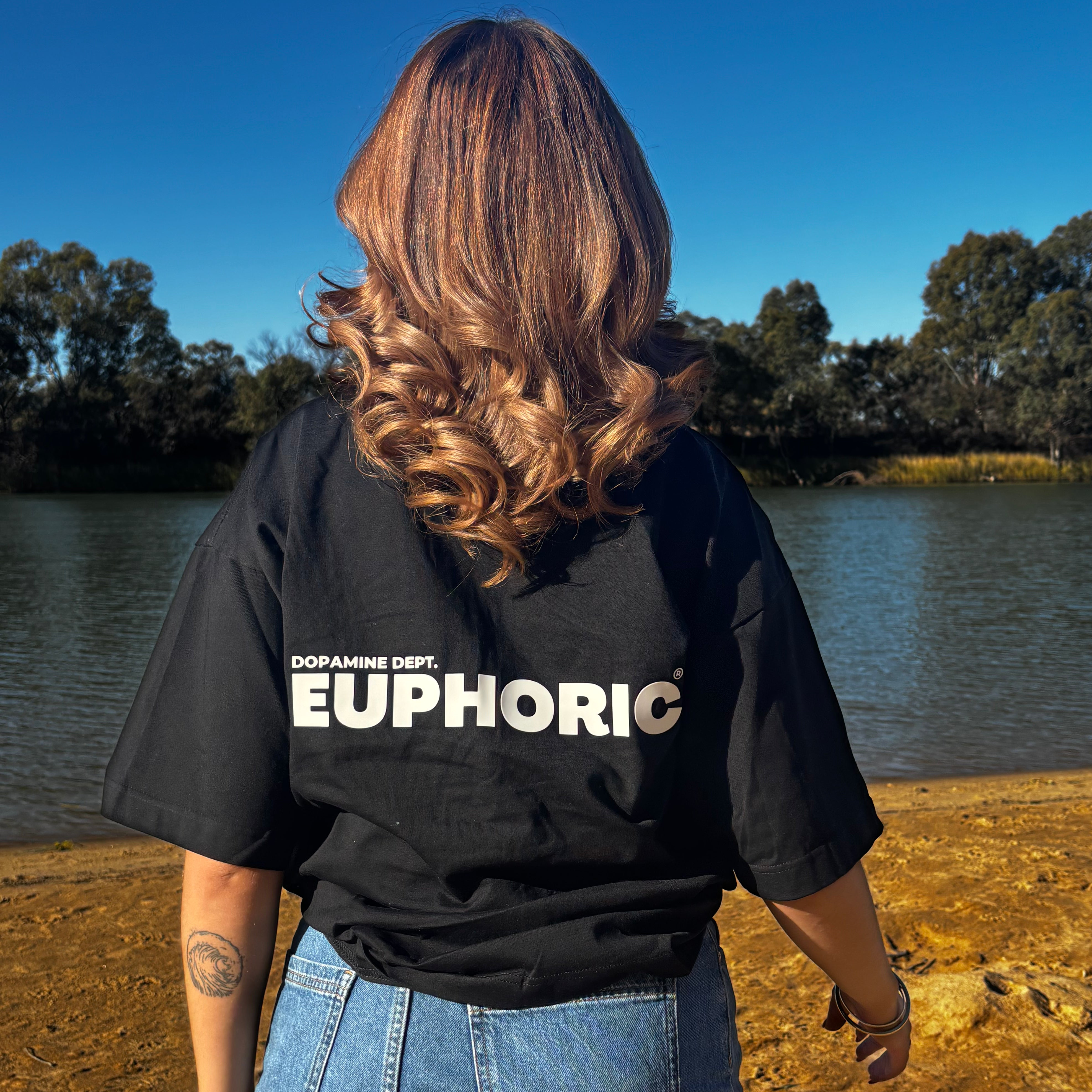 Person wearing a euphoric black t-shirt showing the back logo by a body of water.