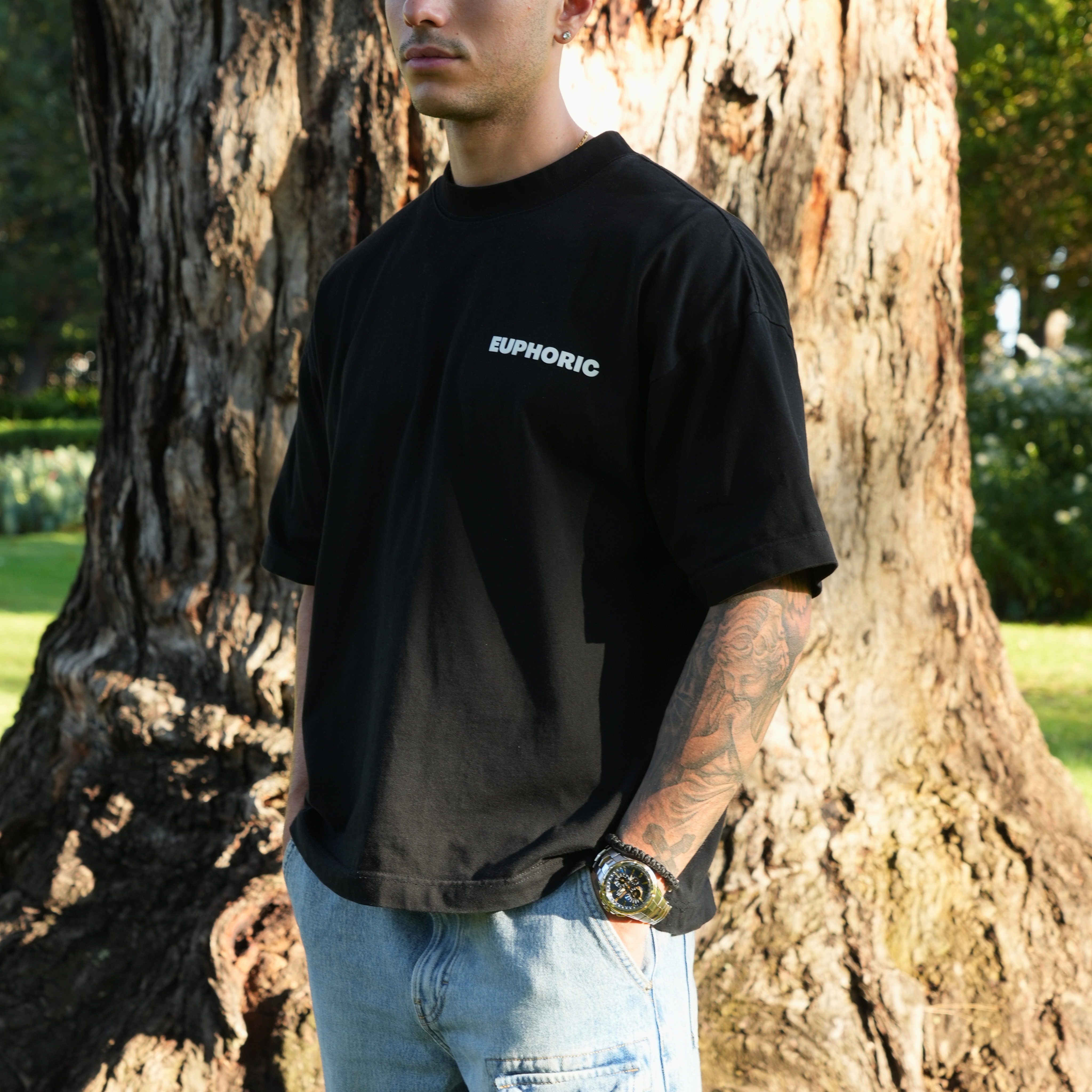 Person wearing a euphoric black t-shirt showing the front of the t-shirt, standing in front of a tree.