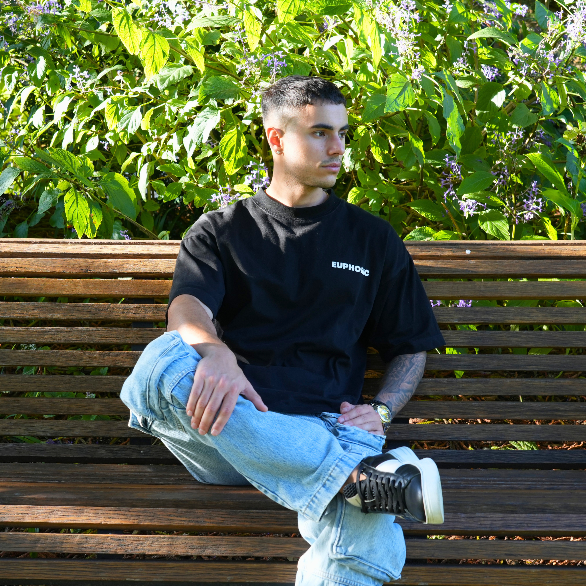 Man sitting on a bench wearing a euphoric black t-shirt and light blue jeans, with green foliage in the background.