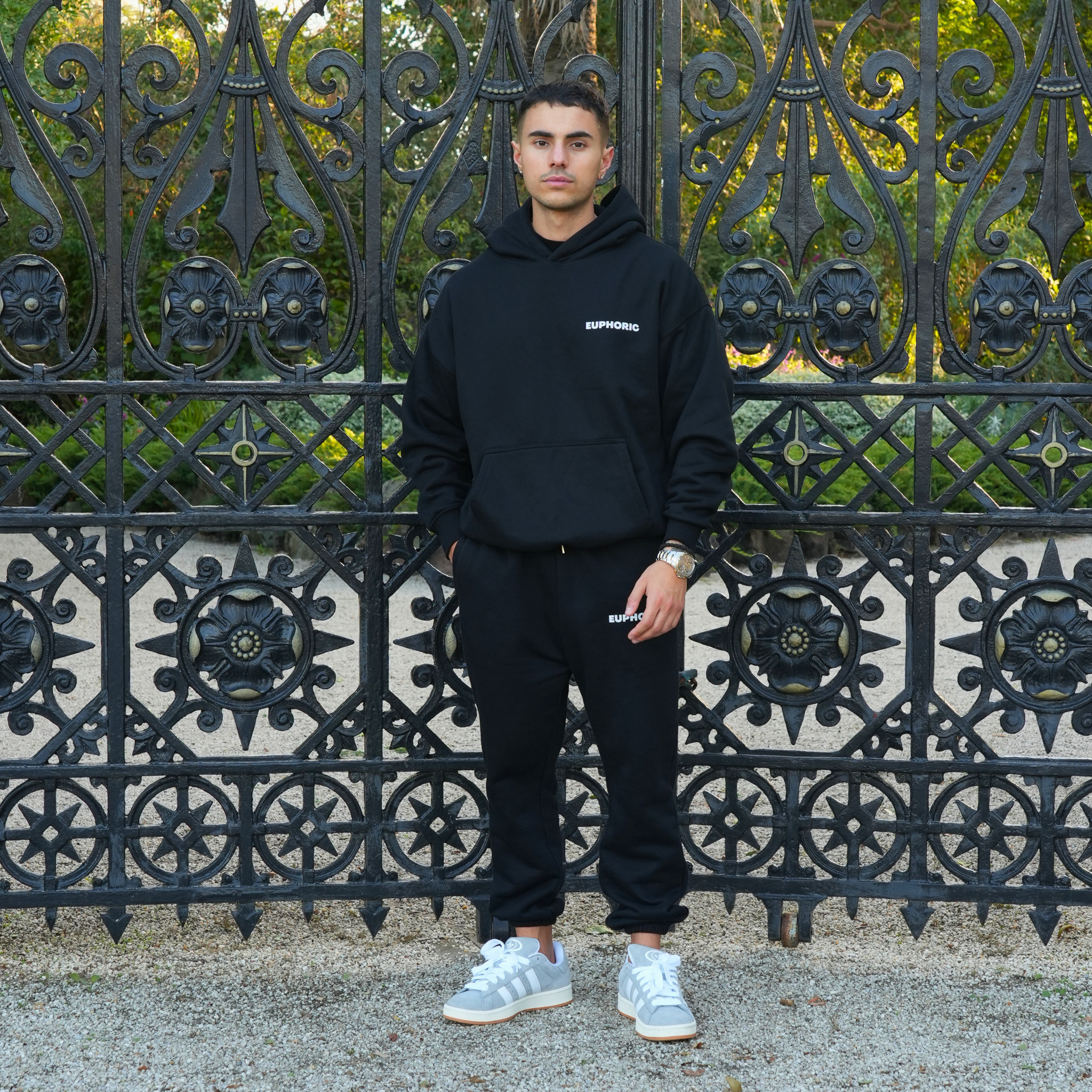 Man wearing a euphoric black hoodie and sweatpants standing in front of an ornate metal gate.