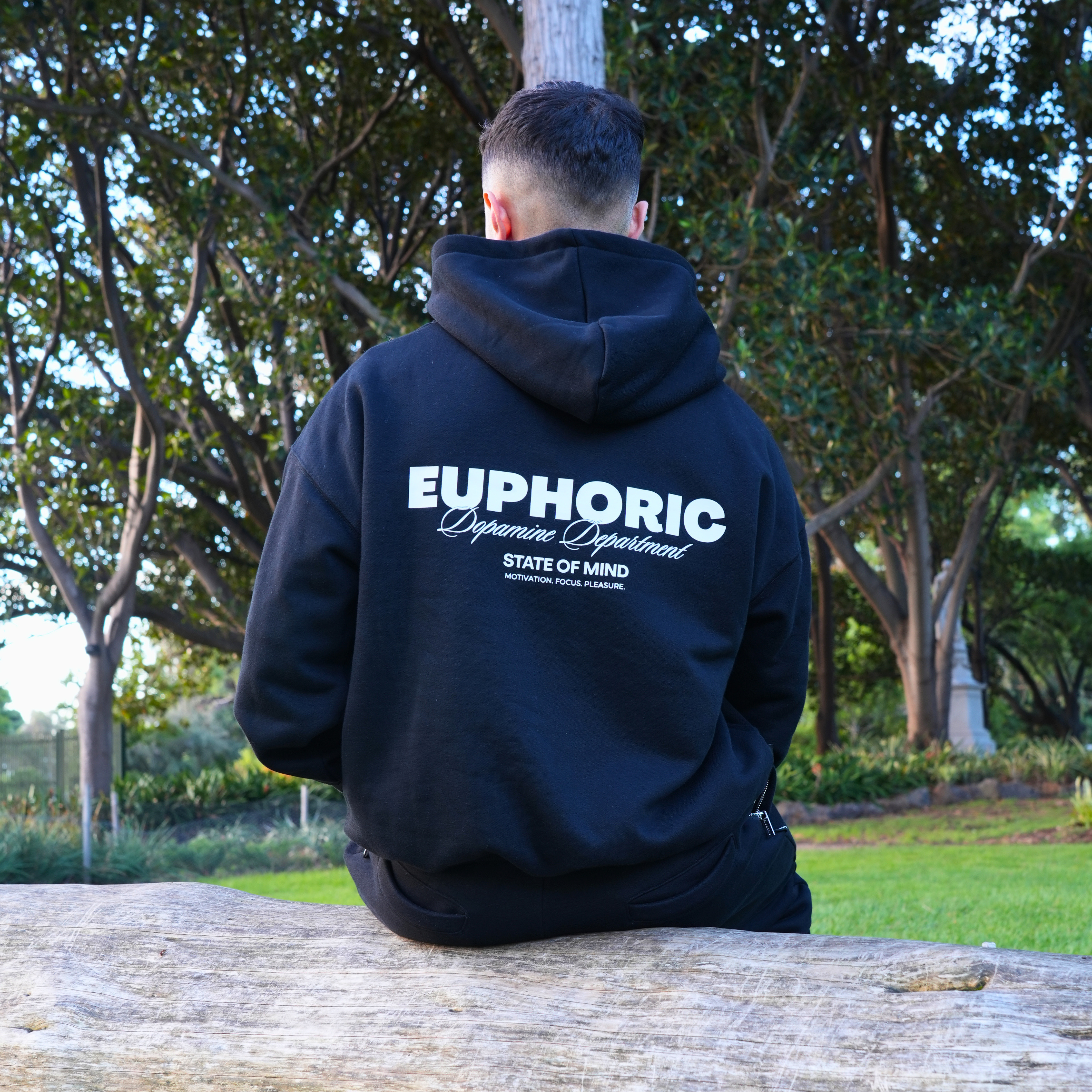 Person wearing a euphoric black hoodie showing the back logo sitting on a log in a park.
