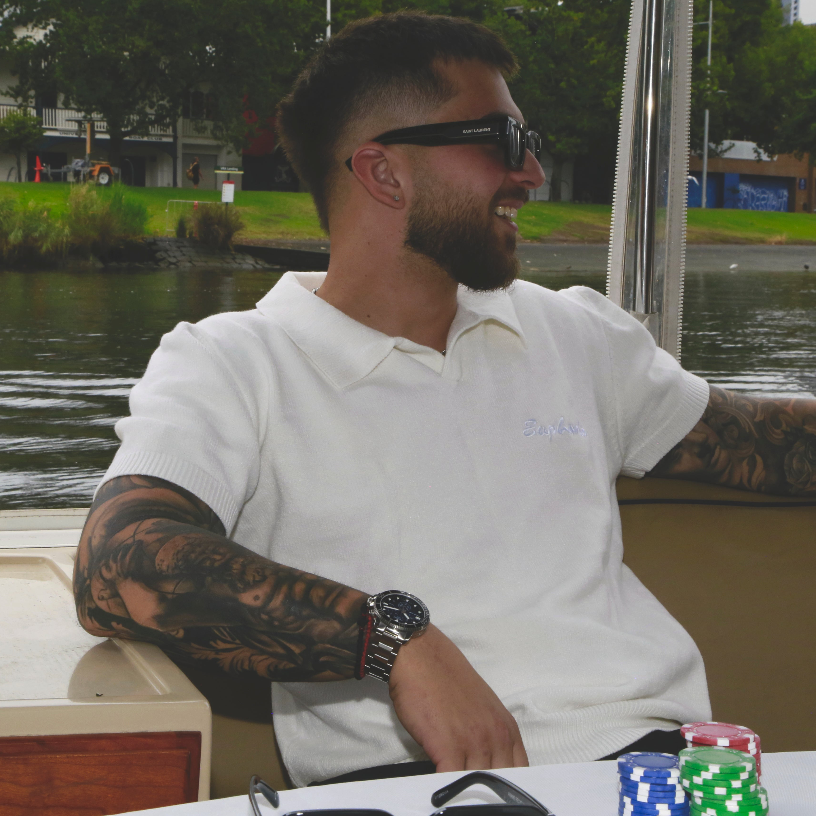 Man wearing a euphoric white polo shirt with tattoos and sunglasses sitting on a boat, smiling.