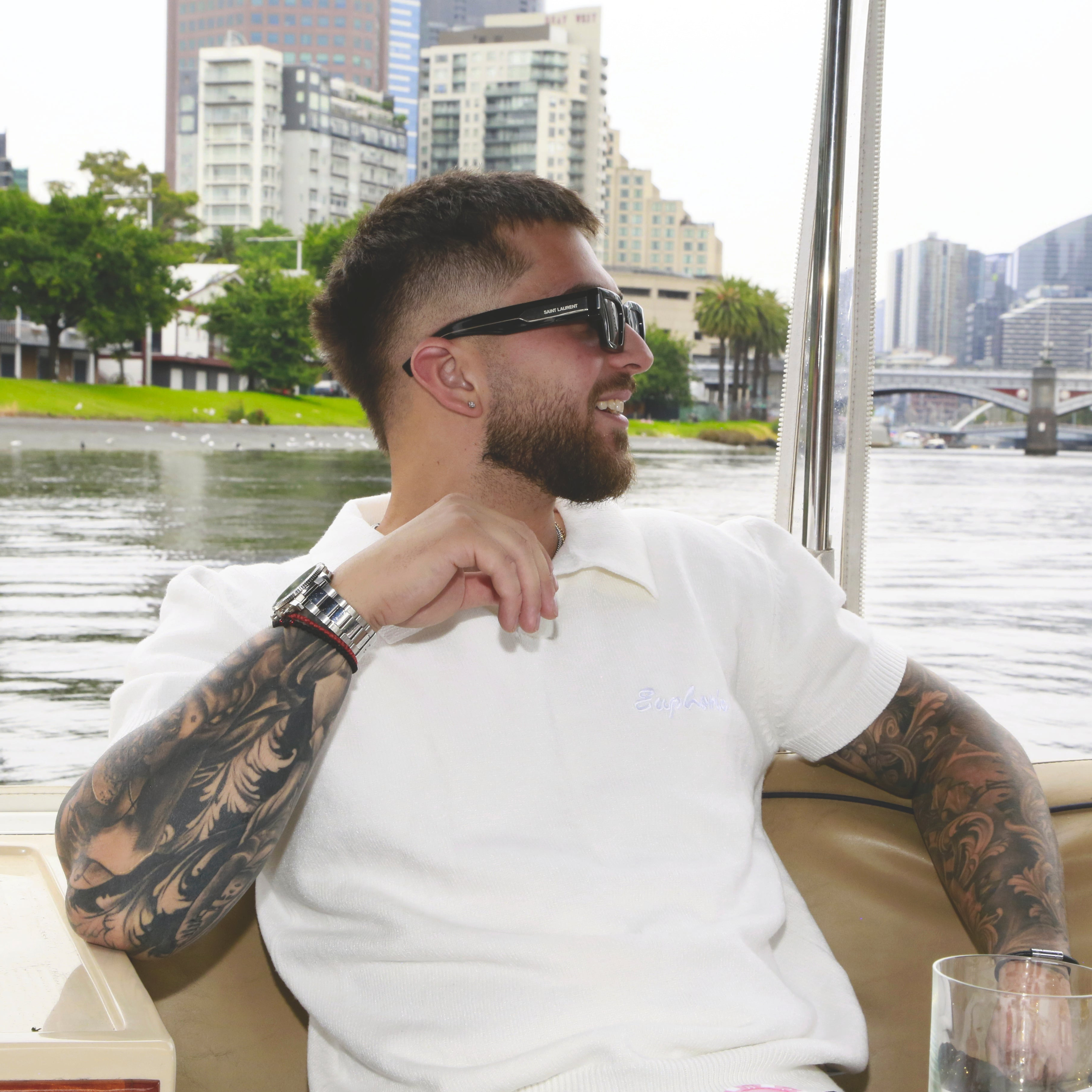 Man wearing euphoric white polo shirt with tattoos wearing sunglasses sitting by a waterfront with cityscape in the background