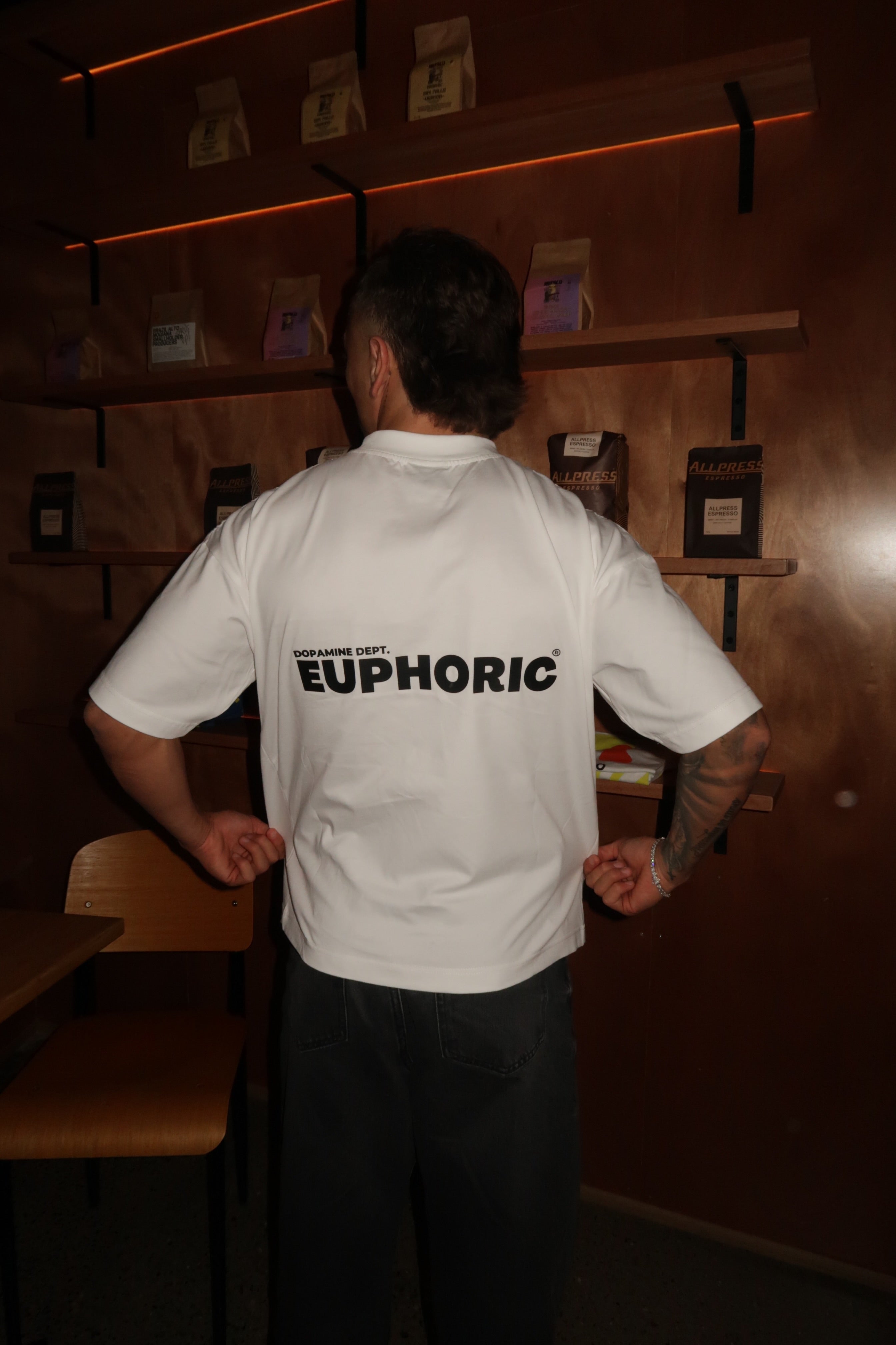 Person wearing a white Euphoric t-shirt in a dimly lit room. Showcasing the back of the t-shirt.