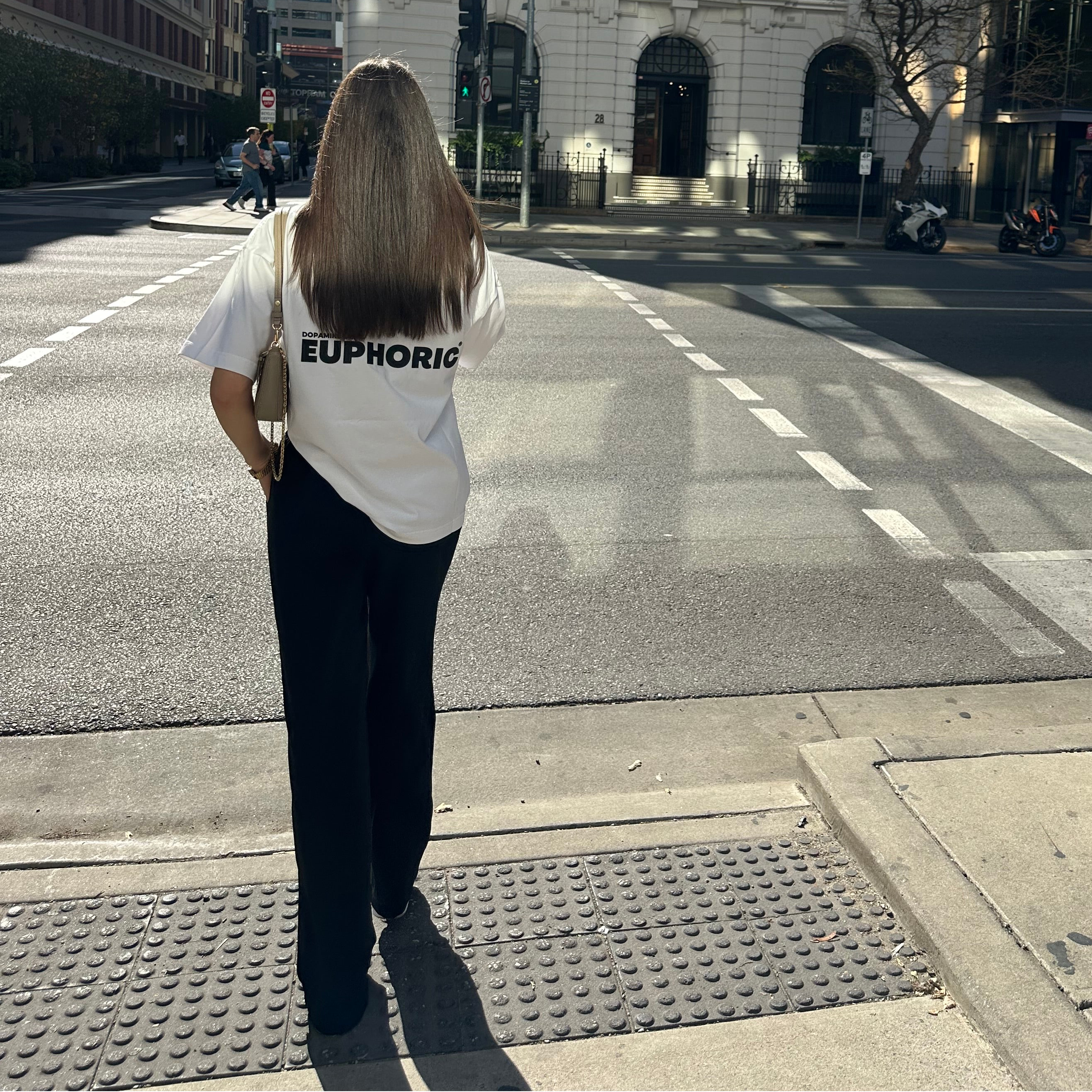 Person wearing a euphoric white t-shirt showing the back of the t-shirt, on a city street.