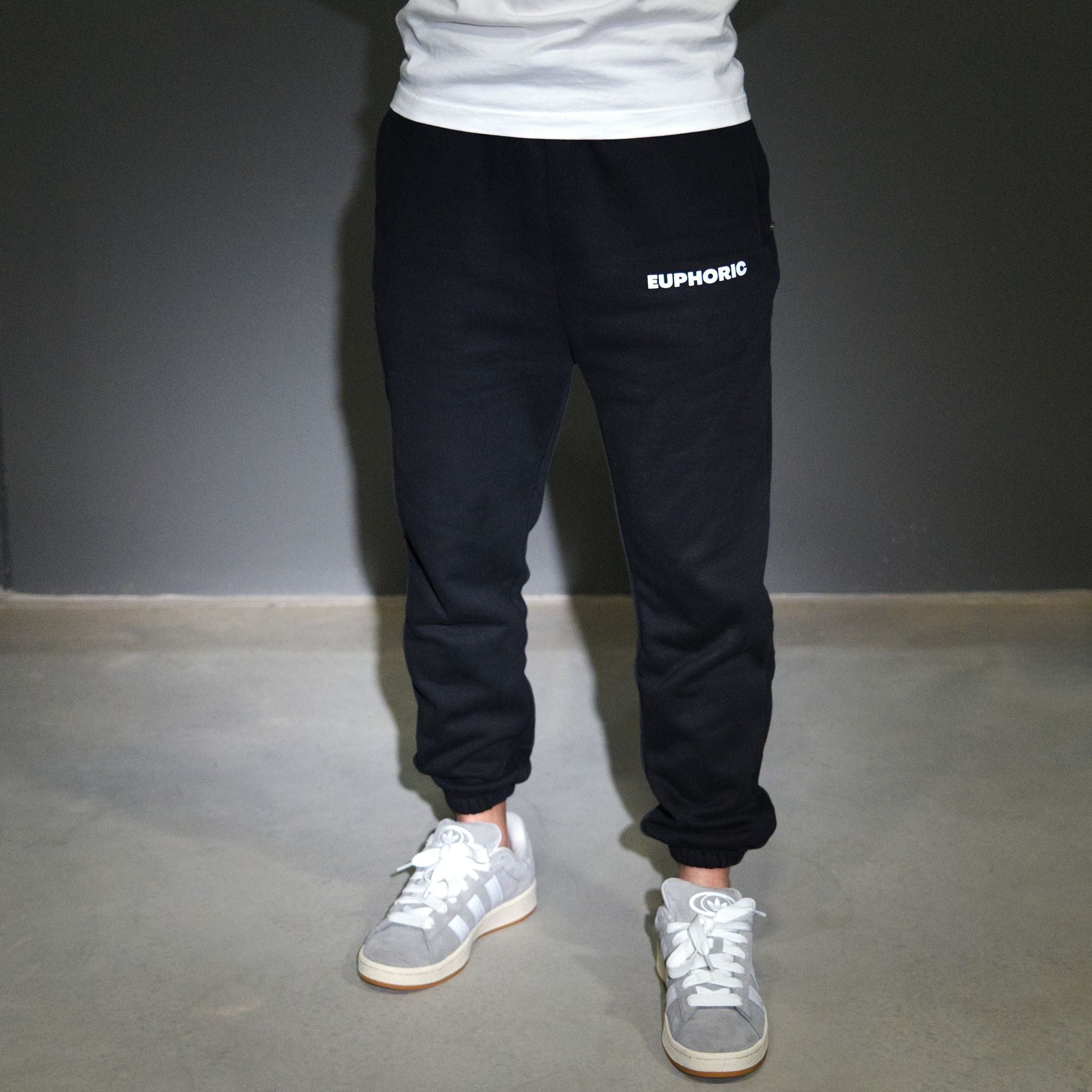 Black jogger sweatpants with 'EUPHORIC' printed in white on the left hip, worn by a model with a white t-shirt and grey sneakers against a grey studio background.