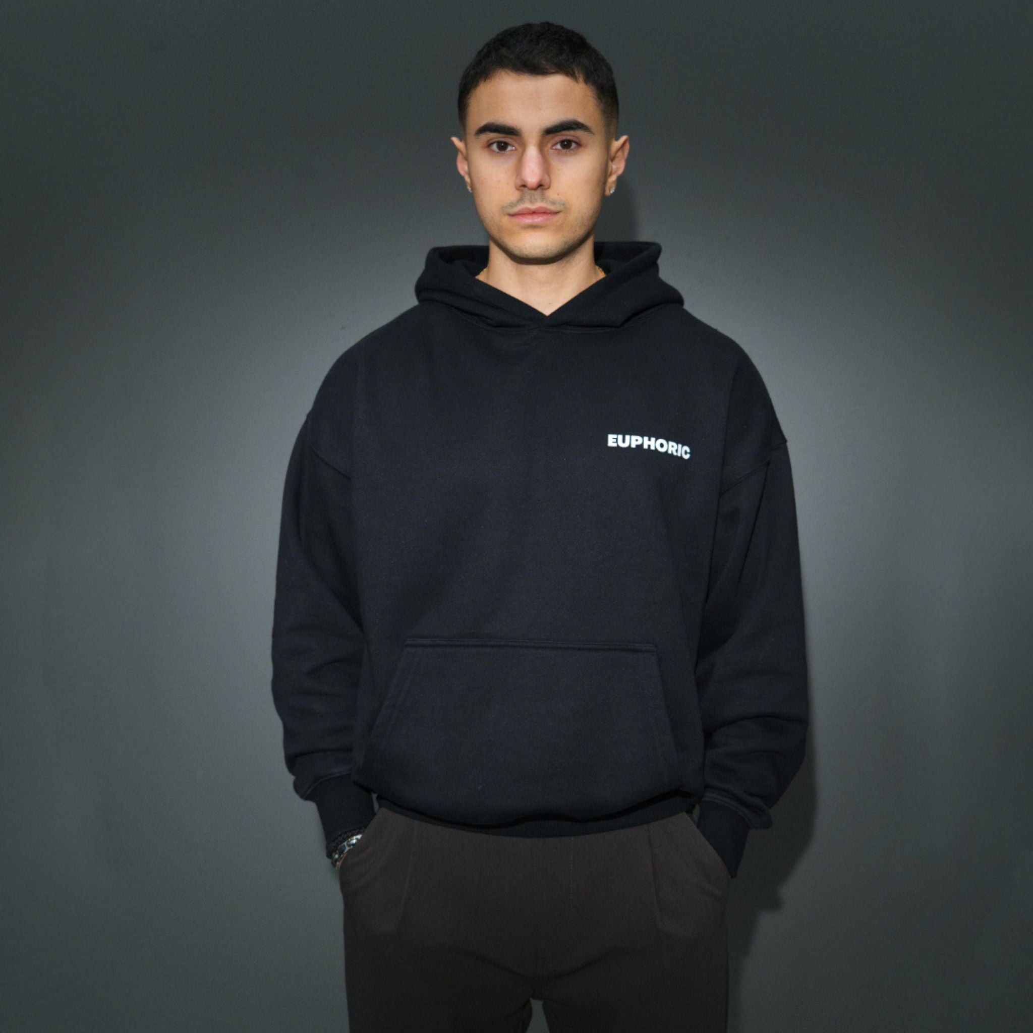 A man wearing a black pullover hoodie with a small white 'EUPHONIC' logo on the left chest, paired with dark brown pants, standing against a dark grey studio background.