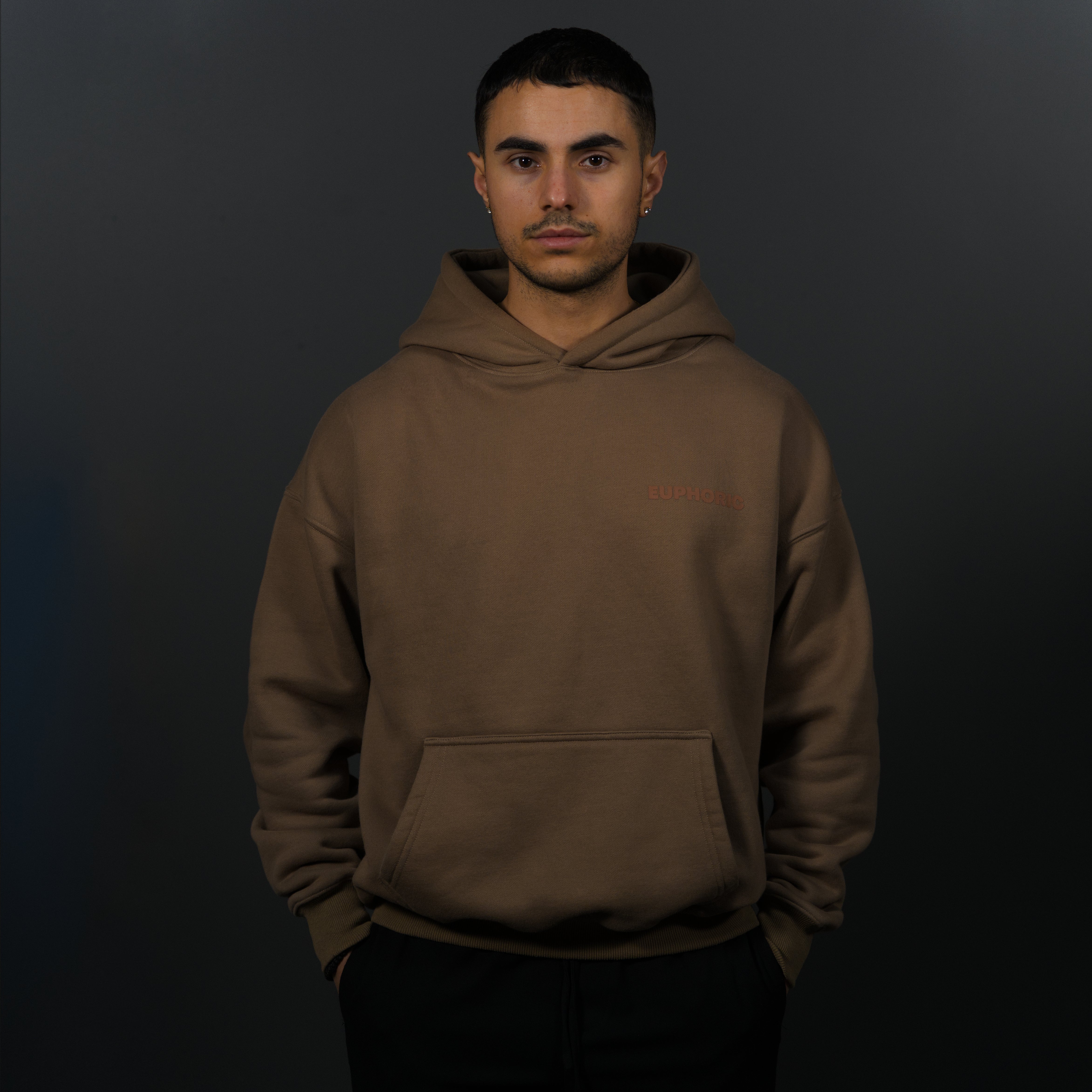 A man wearing a mocha hoodie with a small logo on the left chest, standing against a dark grey background.