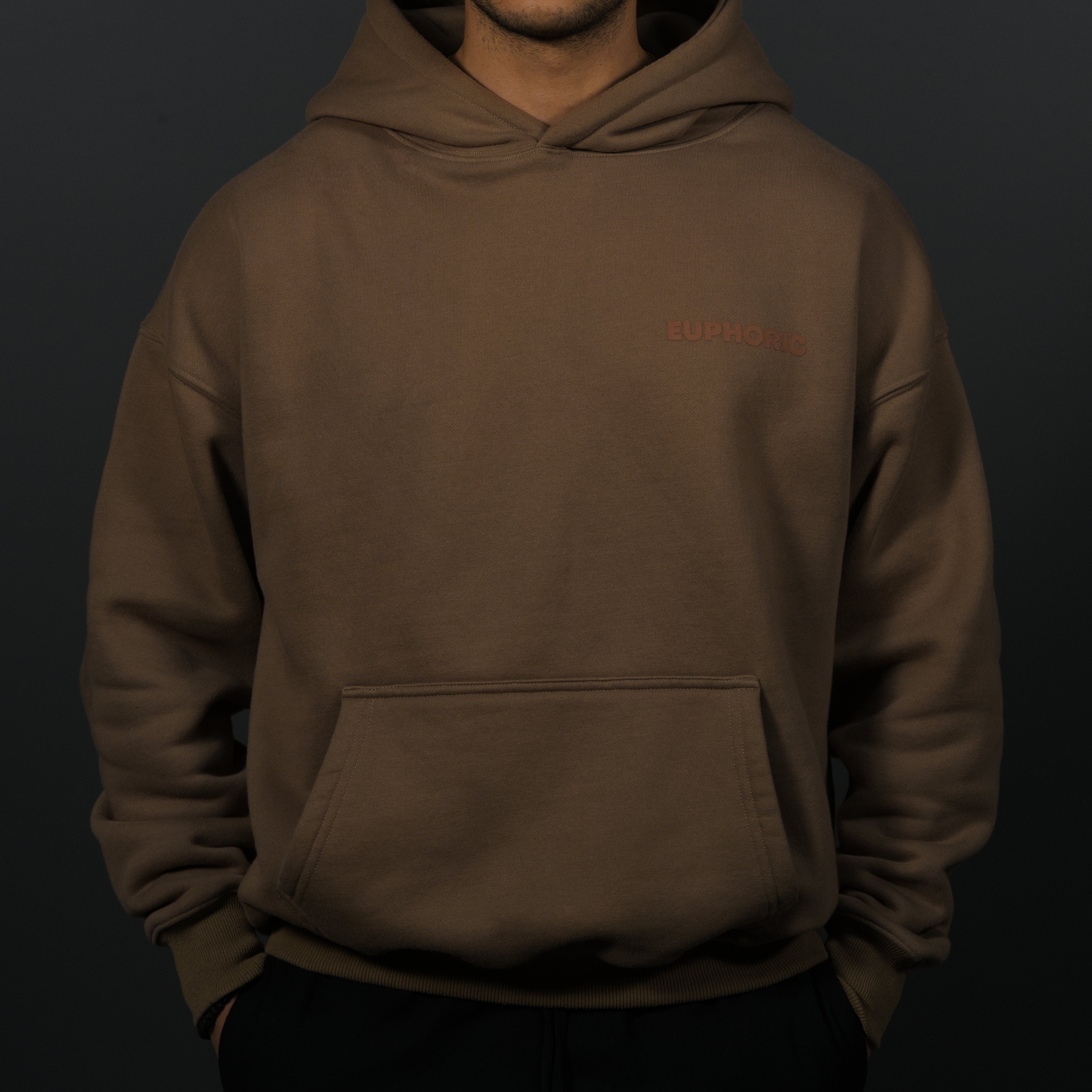 A person wearing a mocha hoodie with a small tonal euphoric logo on the left chest, photographed against a dark studio background.