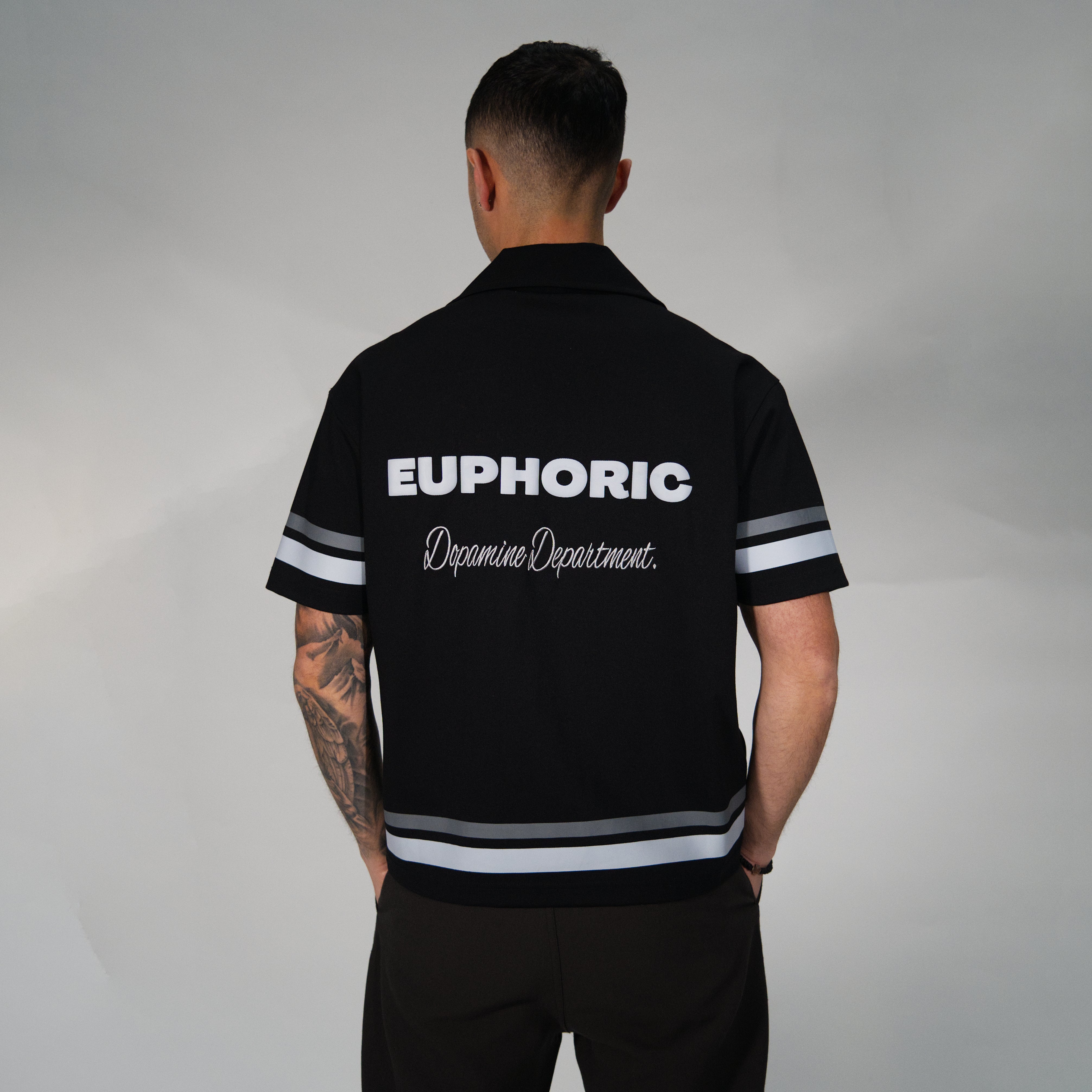 A man viewed from behind wearing a black short-sleeve collared shirt with white and grey striped accents on the sleeves and hem, featuring 'EUPHORIC' and 'Dopamine Department.' printed in white across the back.
