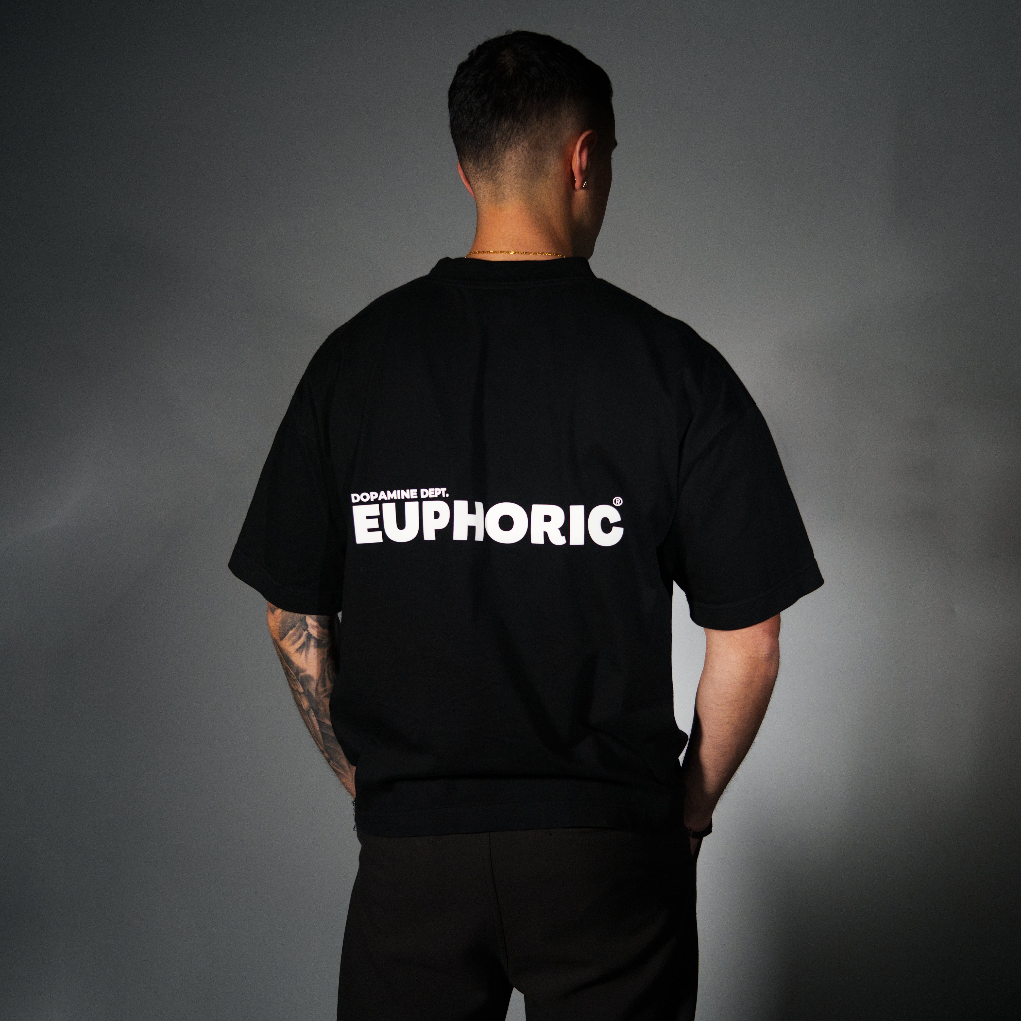 A man viewed from behind wearing a black t-shirt with 'EUPHORIC' printed in bold white letters on the back, set against a dark grey studio background.