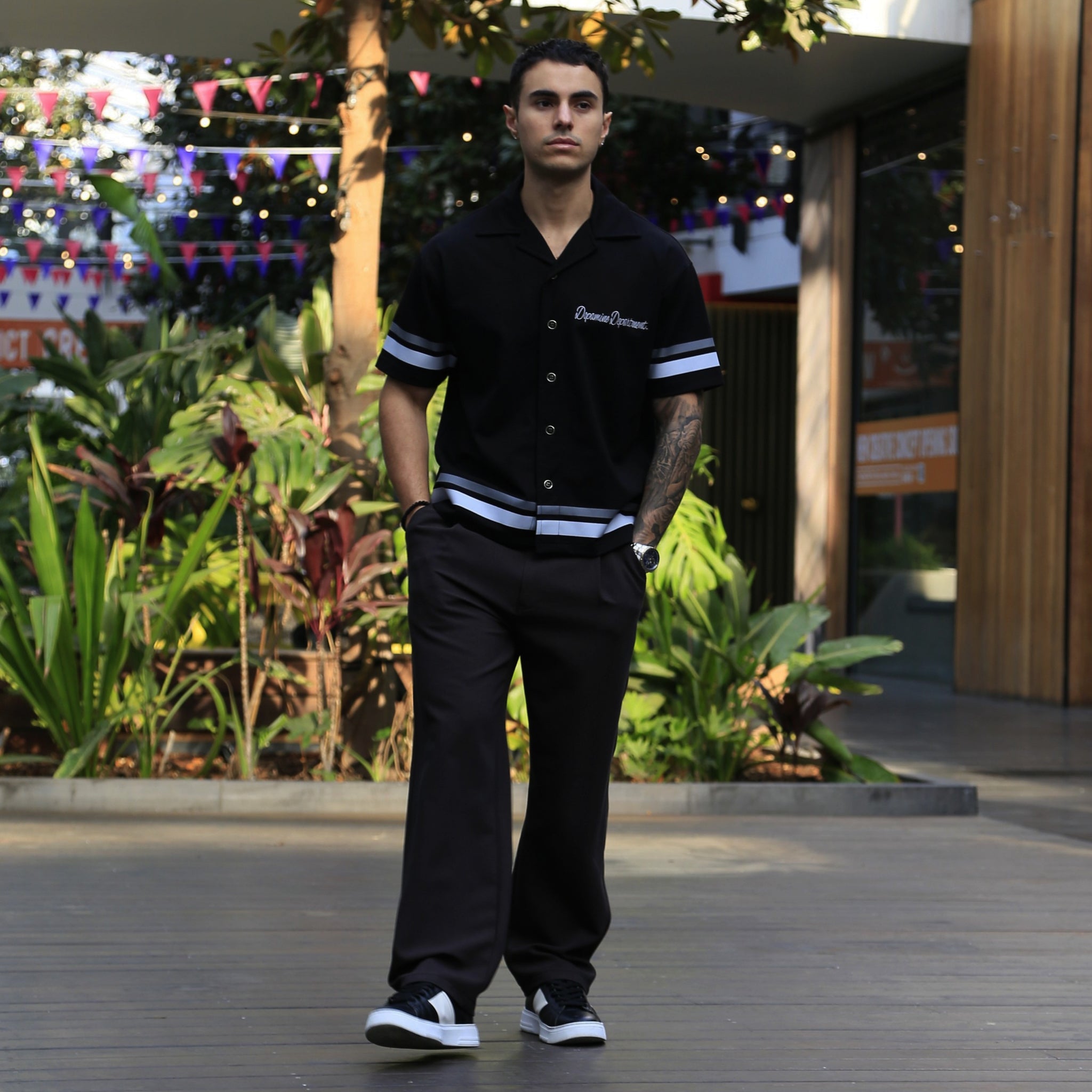 Man wearing a Euphoric Black Relaxed Button Shirt standing outdoors with plants and decorations in the background