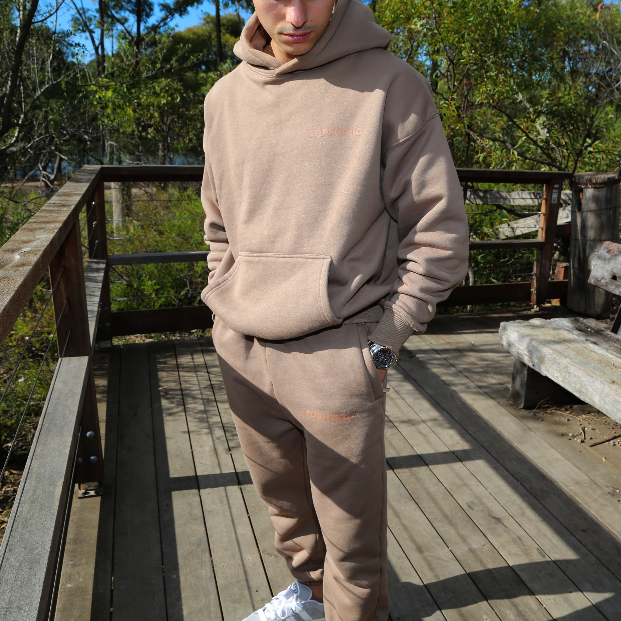Person wearing a euphoric essential mocha tracksuit set on a wooden deck with trees in the background