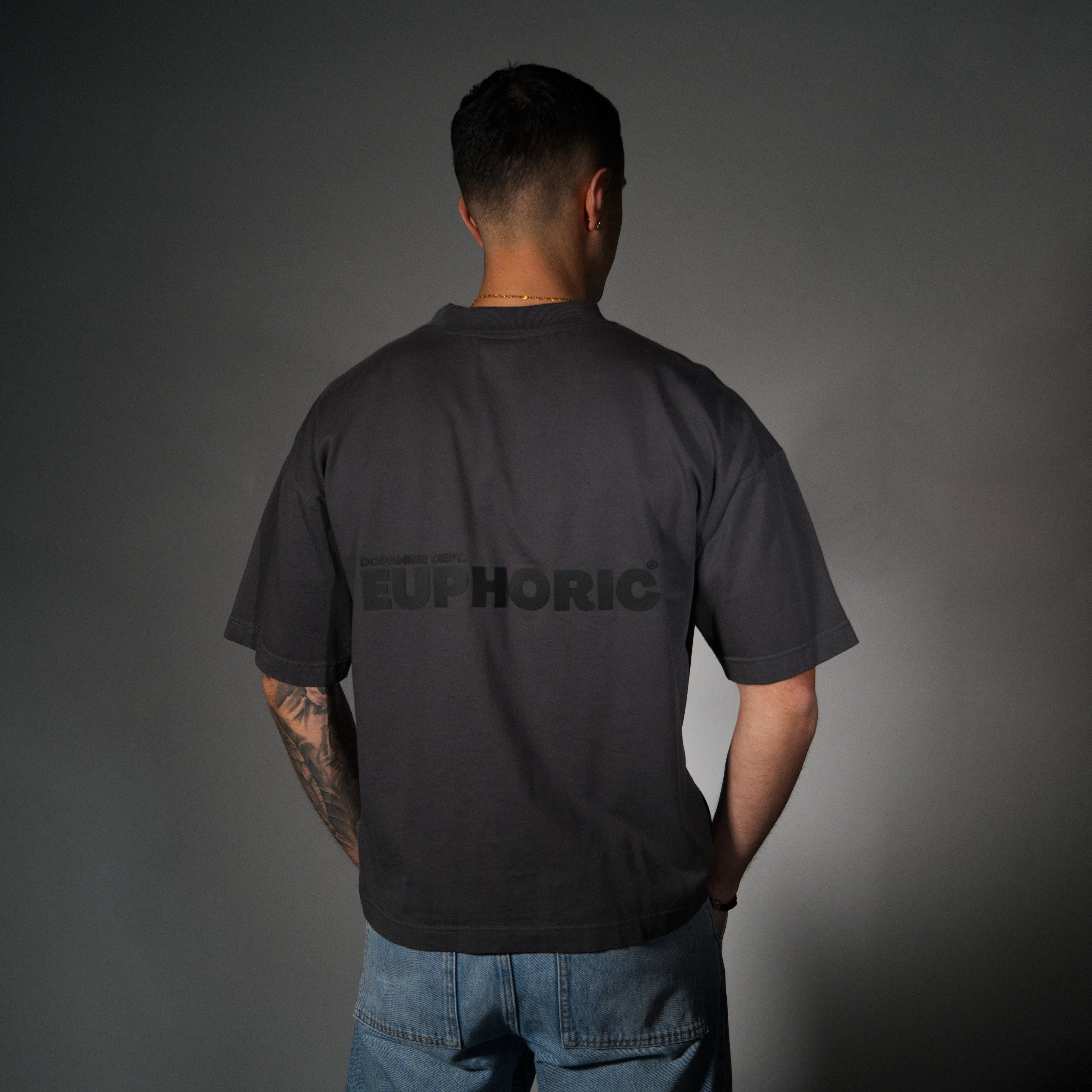 A man viewed from behind wearing a dark charcoal grey t-shirt with 'EUPHORIC' printed in tonal black across the back, paired with blue jeans against a dark grey studio background.