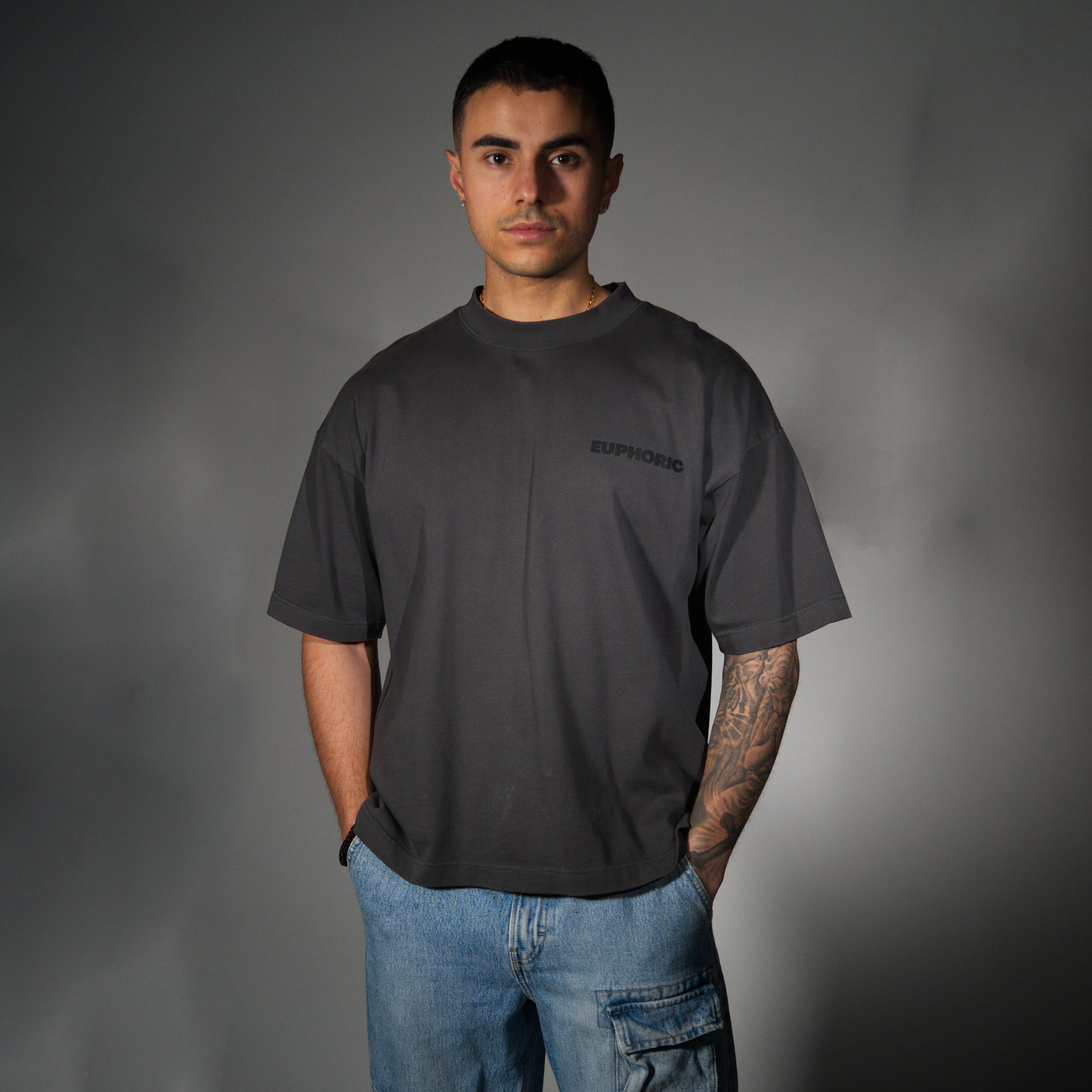 A man wearing a dark grey oversized t-shirt with a subtle black 'EUPHORIC' logo on the left chest, paired with light blue jeans, standing against a dark grey studio background.