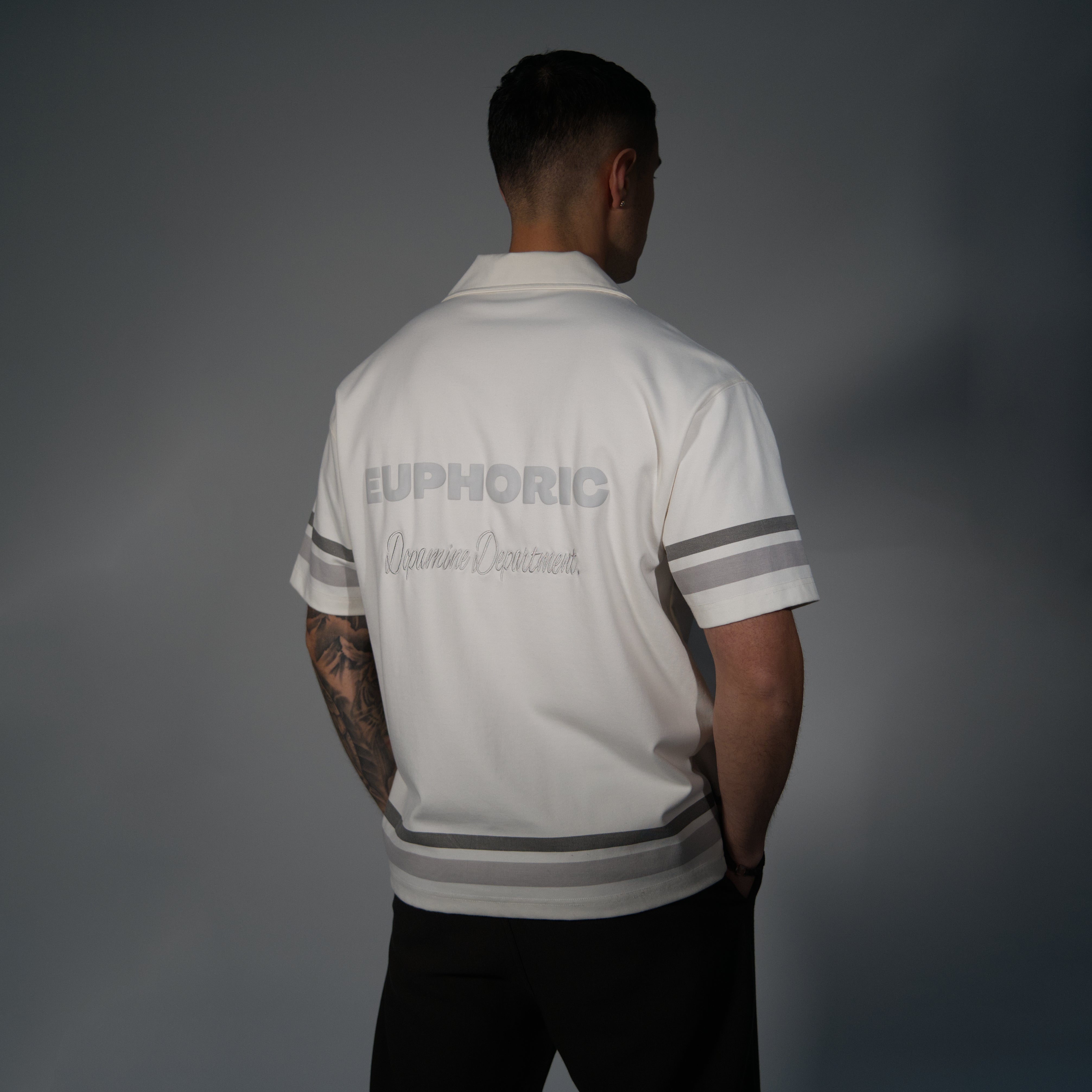 A man viewed from behind wearing a white short-sleeve polo shirt with grey stripes on the sleeves and hem, and grey text on the back reading 'EUPHORIC' and 'Dopamine Department.' against a dark grey studio background.