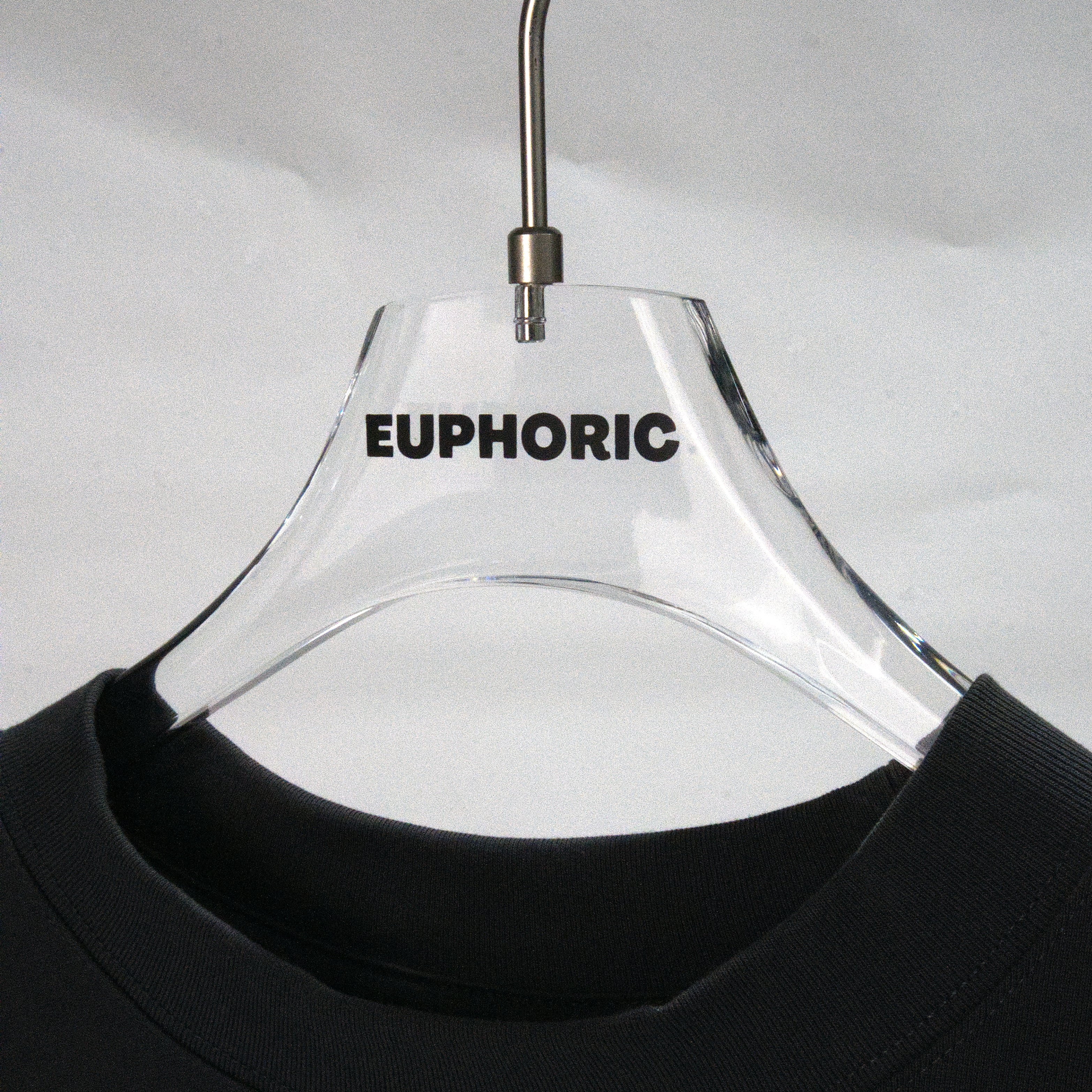 A clear acrylic hanger with the word 'EUPHORIC' printed in black on the front, holding the collar of a black crewneck shirt against a light gray background.