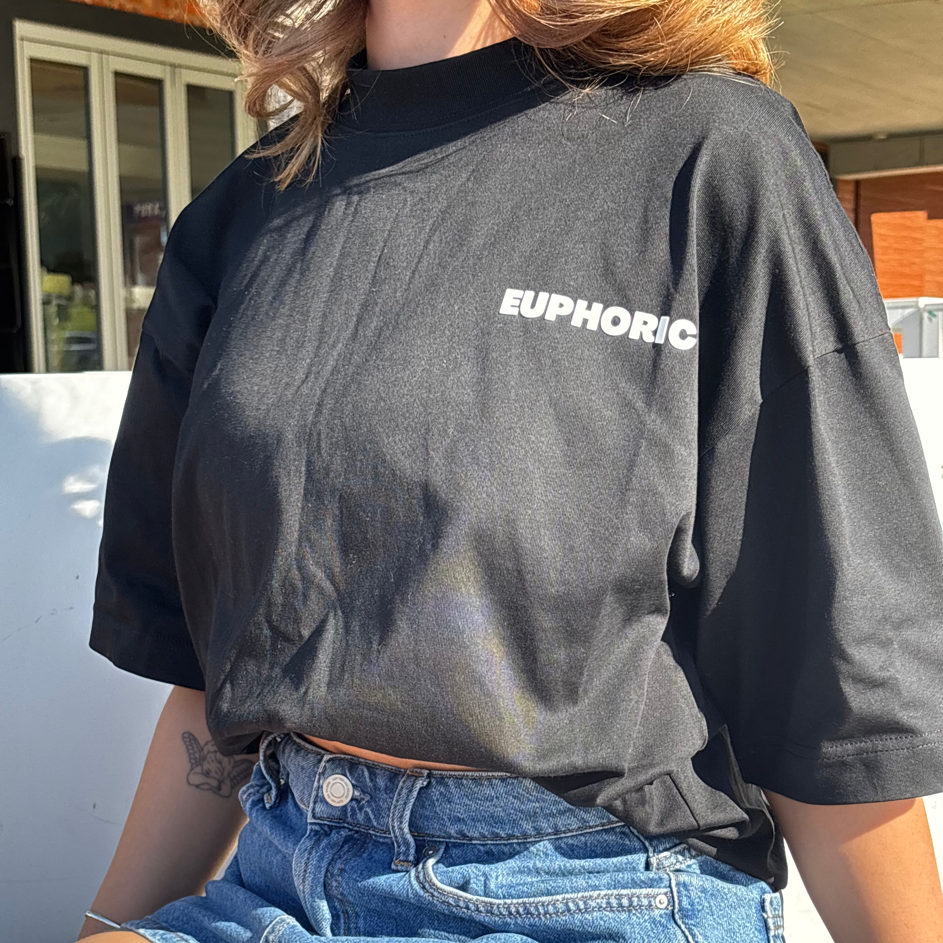 Close-up of person wearing a euphoric black t-shirt showing front logo, sitting outdoors.