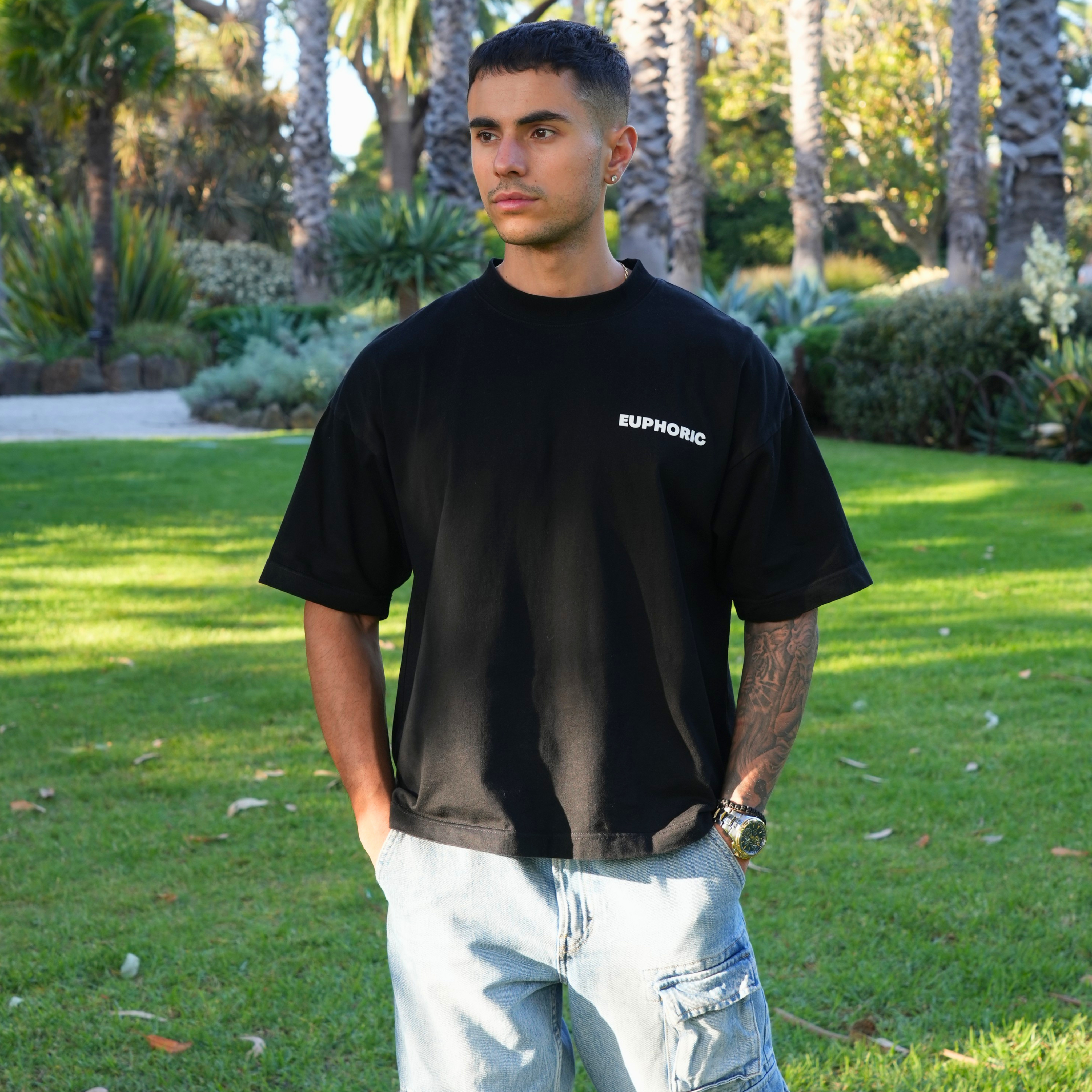 Man wearing euphoric black t-shirt showing the front of the t-shirt, standing outdoors in a garden.