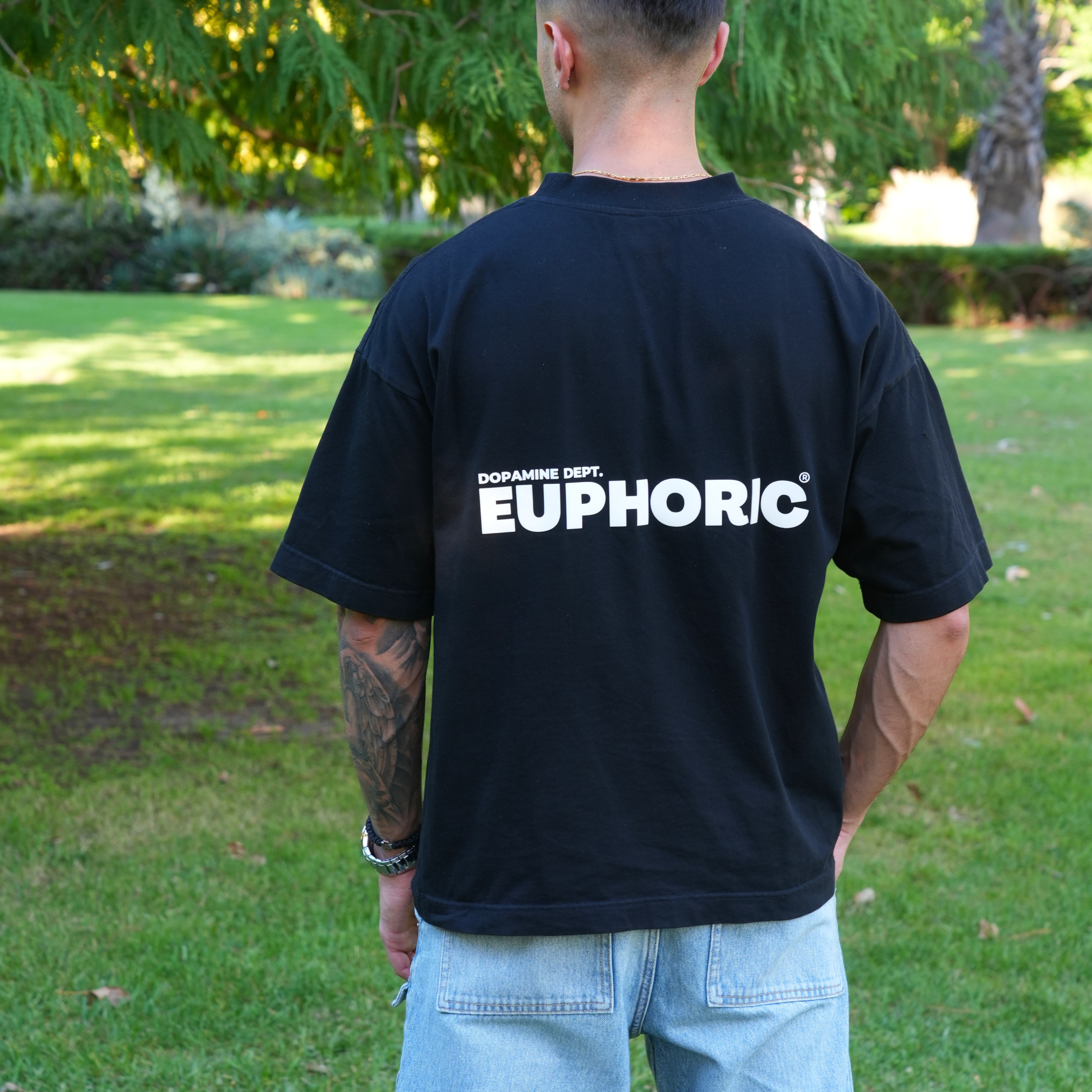 Person wearing a euphoric black t-shirt showing the back of the t-shirt logo in an outdoor setting