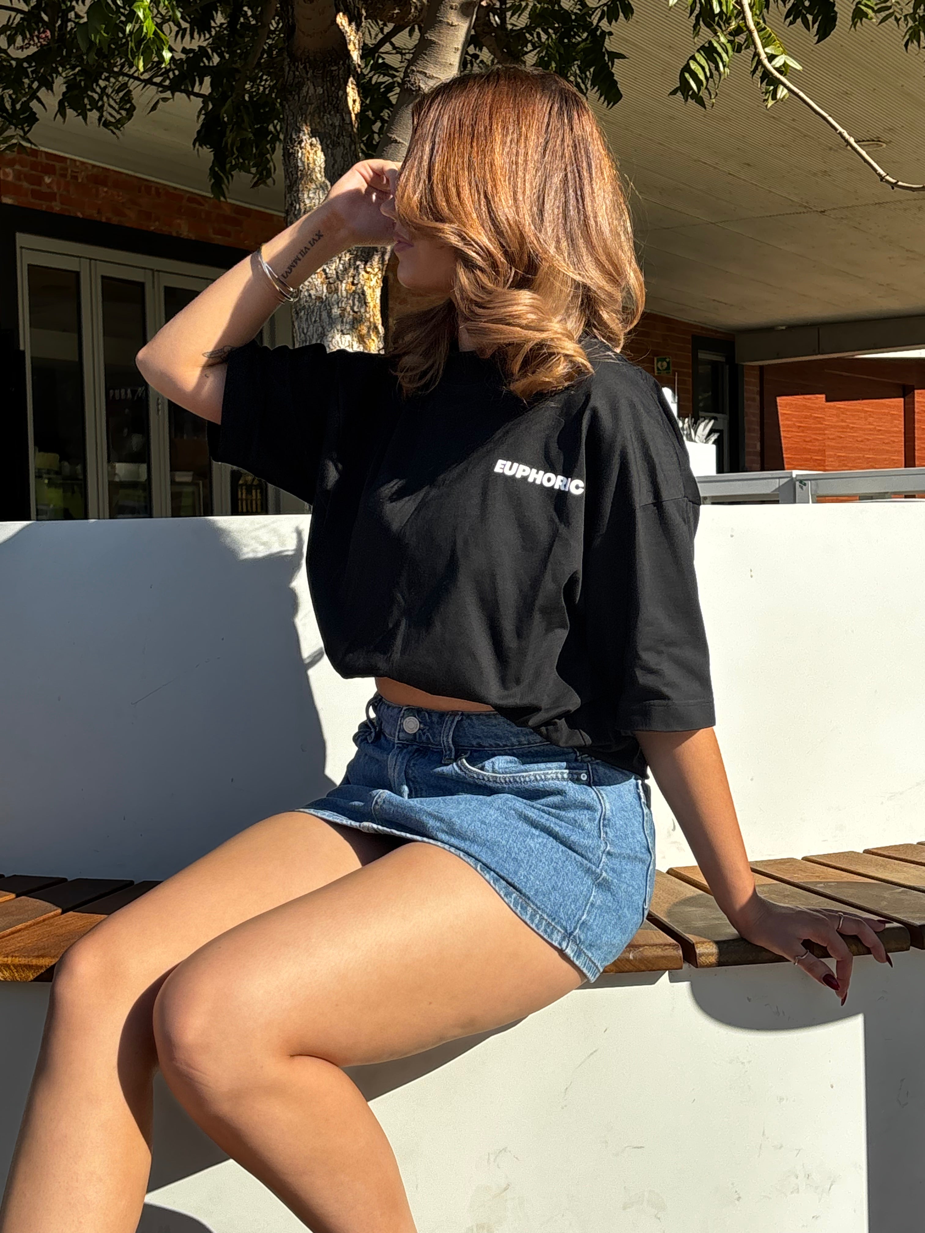 Person wearing a black Euphoric T-Shirt and denim shorts sitting outdoors.