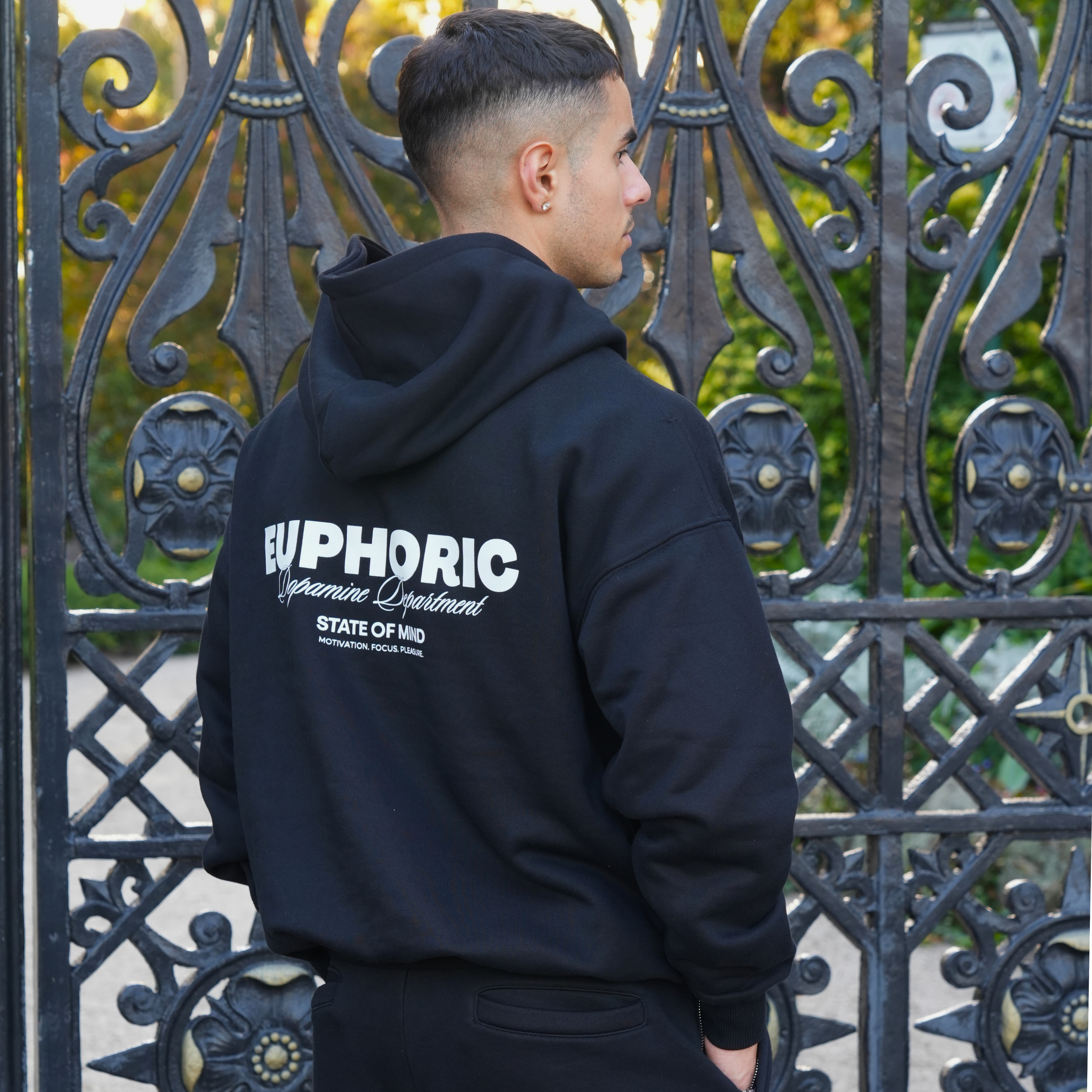 Person wearing a euphoric black hoodie showcasing the logo on the back of the hoodie, standing in front of an ornate metal gate.