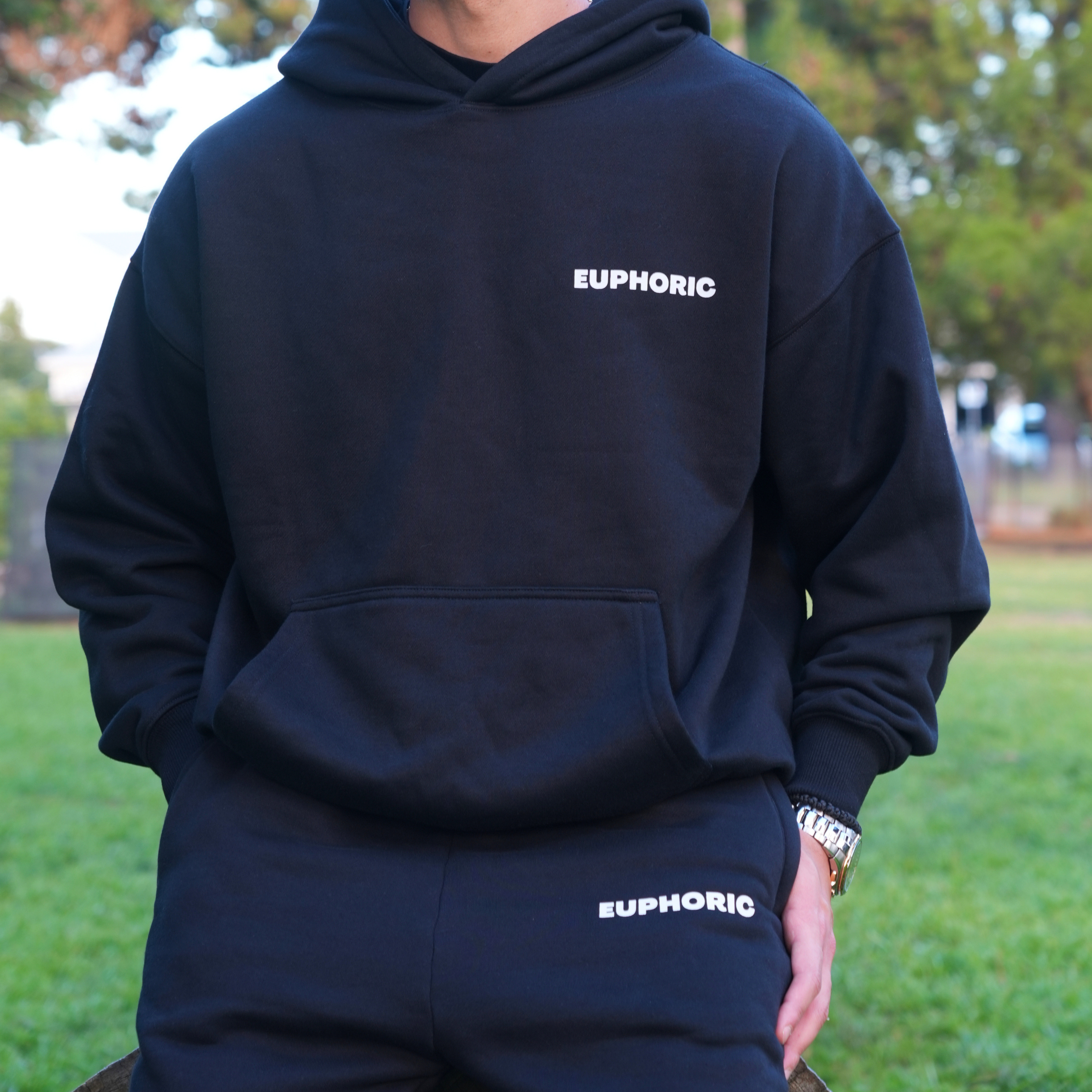 Person wearing a black hoodie and pants with 'EUPHORIC' text, standing outdoors.