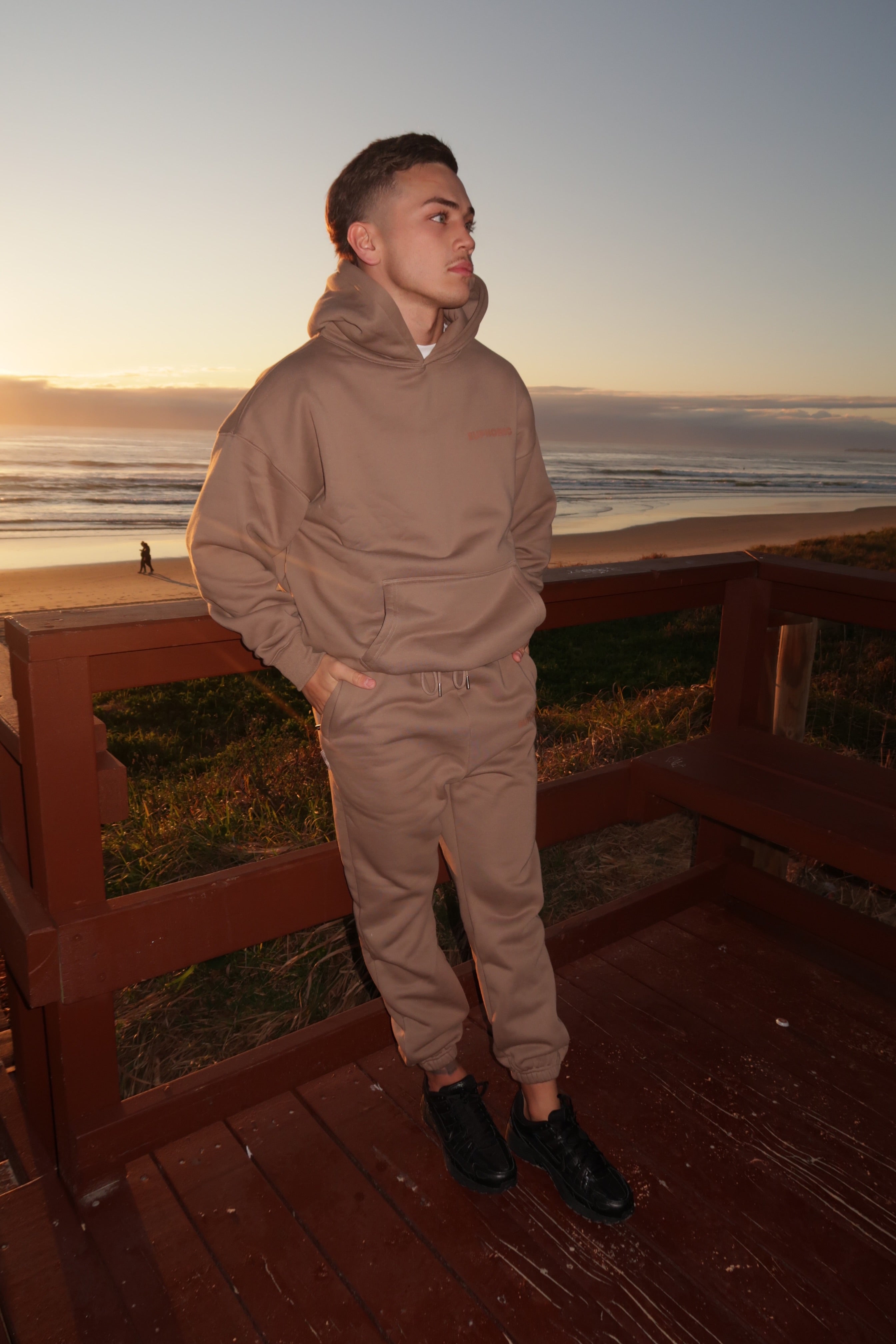 Man in Euphoric mocha tracksuit standing on a wooden platform by the beach at sunset.
