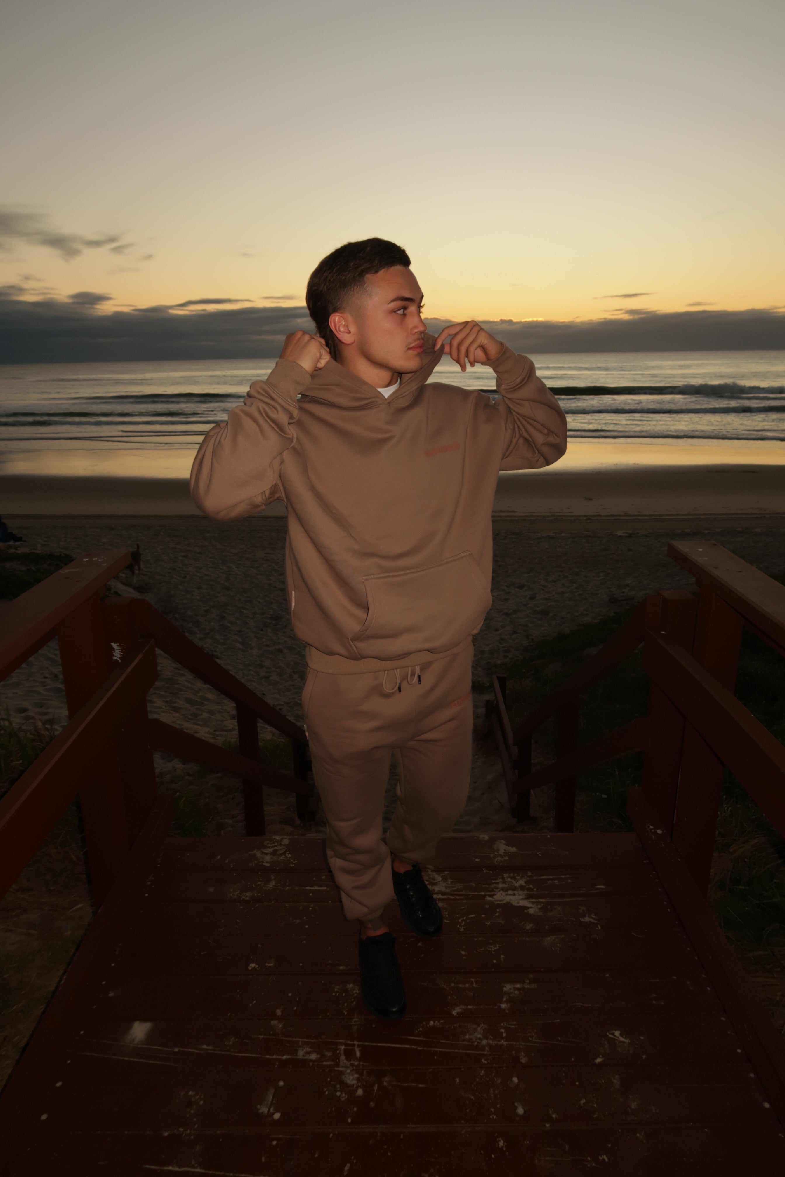 Man in Euphoric mocha tracksuit standing on a wooden platform by the beach at sunset.