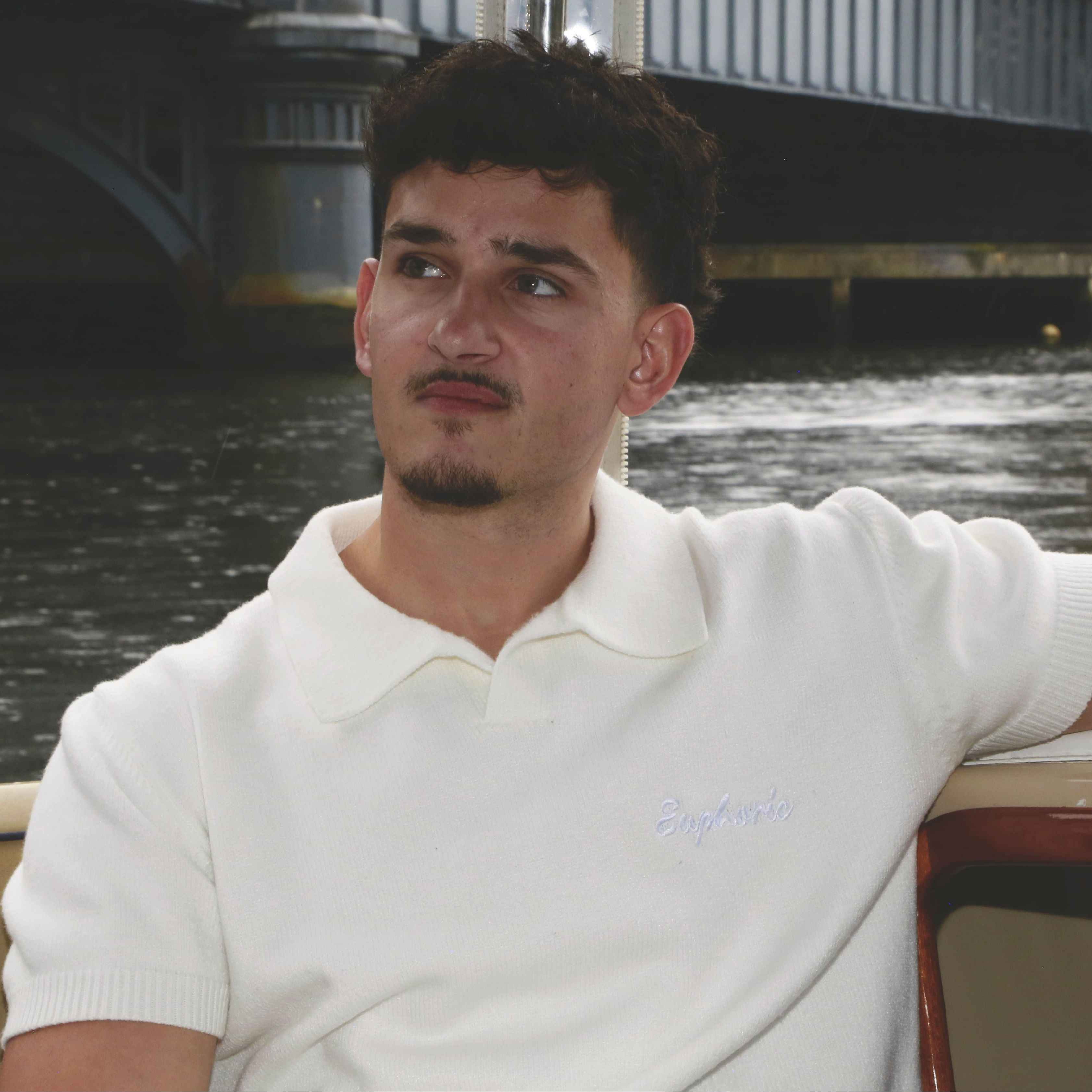Man in euphoric white polo shirt with a logo sitting by a waterfront with a bridge in the background
