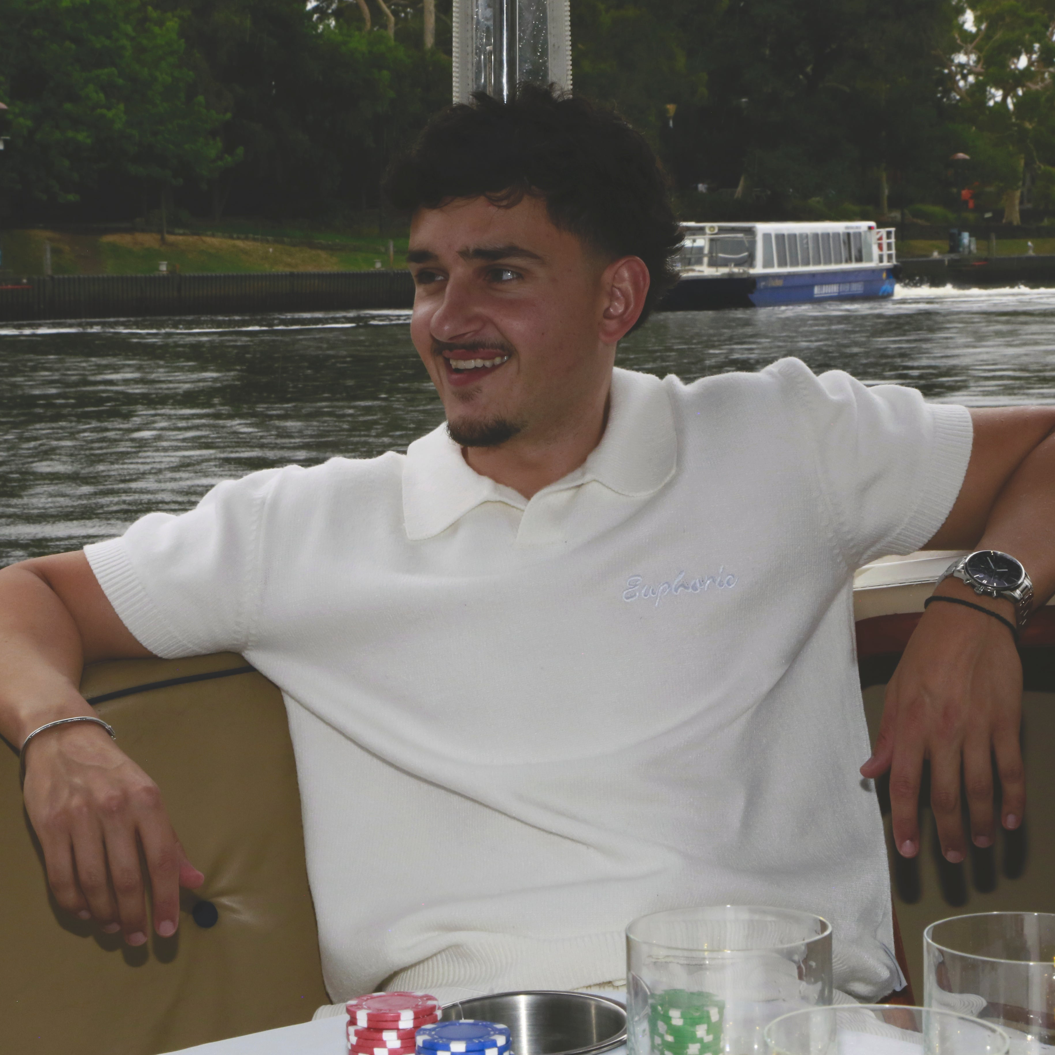 Man in a euphoric white polo shirt sitting by a body of water with a boat in the background