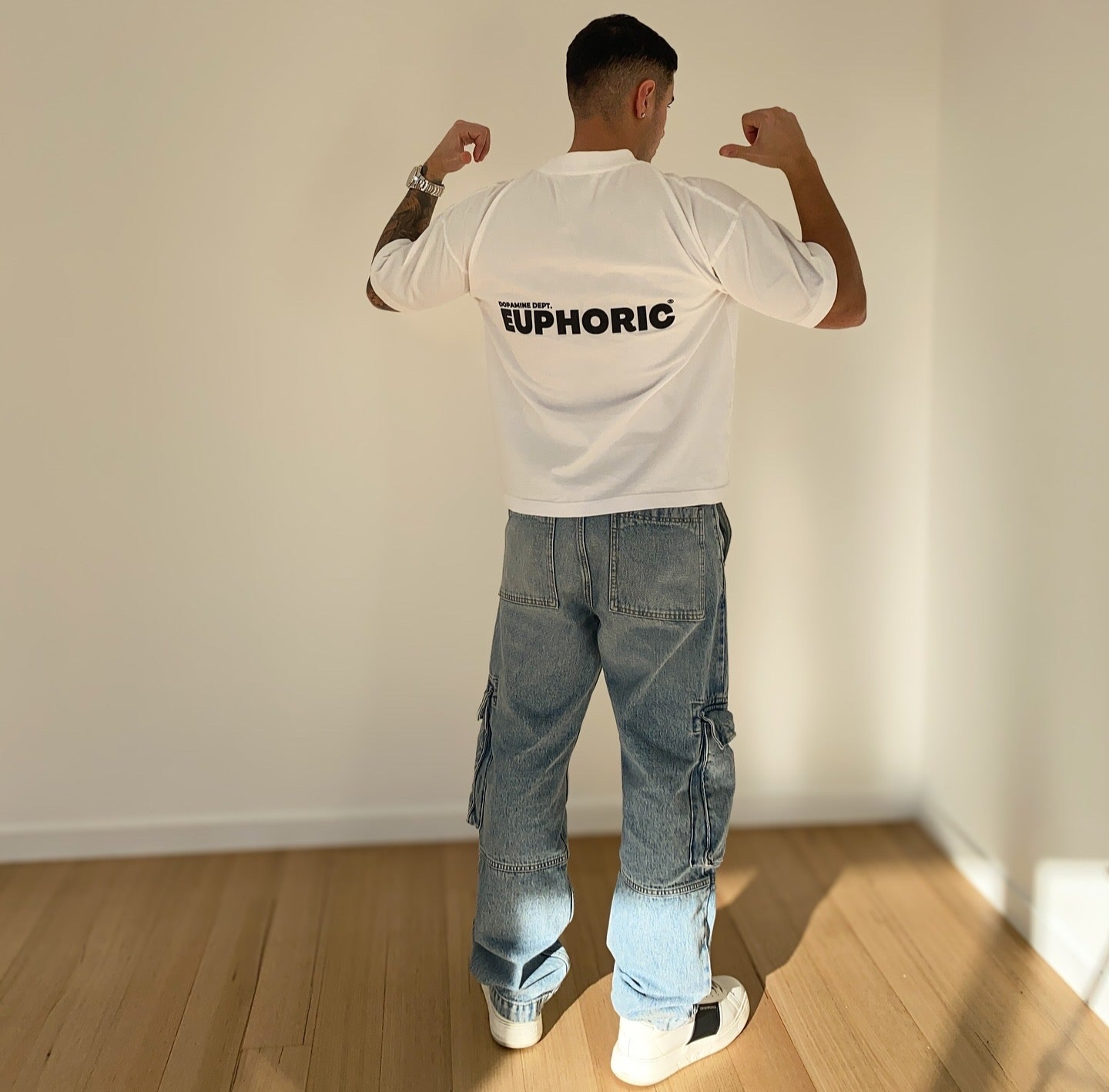Person wearing a euphoric white t-shirt and blue jeans, standing in a room with wooden floor and white walls.