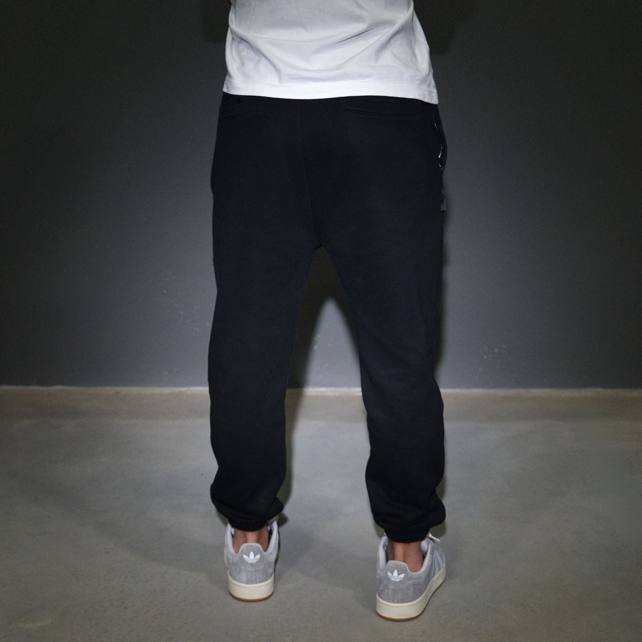 A person shown from the waist down wearing dark navy jogger sweatpants, a white t-shirt, and light grey sneakers, standing against a dark grey wall.