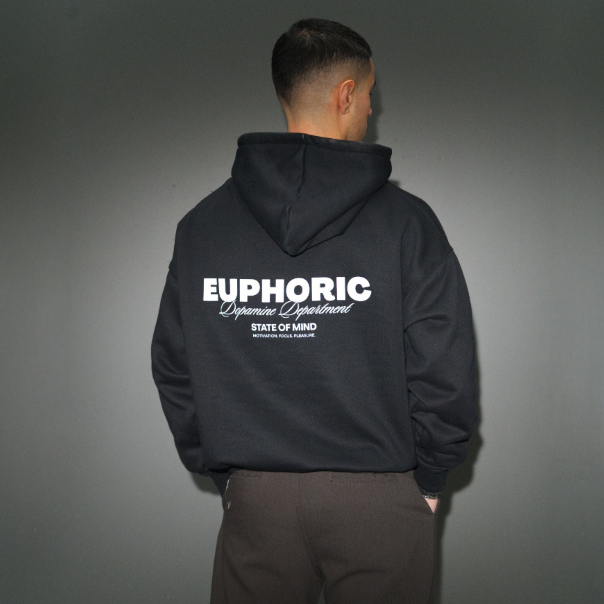 A man viewed from behind wearing a black hoodie with 'EUPHORIC Department' printed in white on the back, paired with dark brown pants, set against a dark grey background.