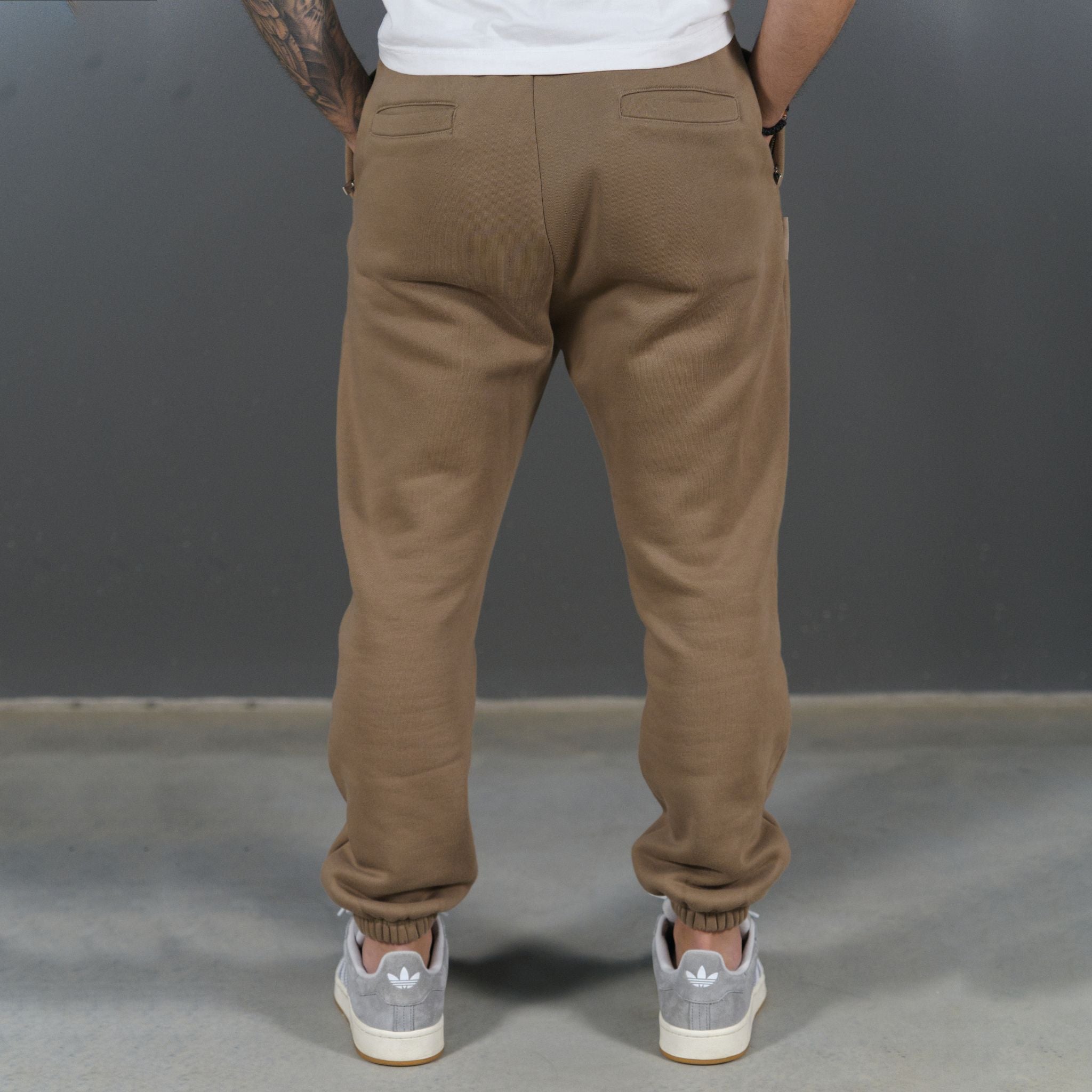 A person shown from the back wearing loose-fit mocha jogger sweatpants, a white t-shirt, and grey sneakers against a dark grey studio background.