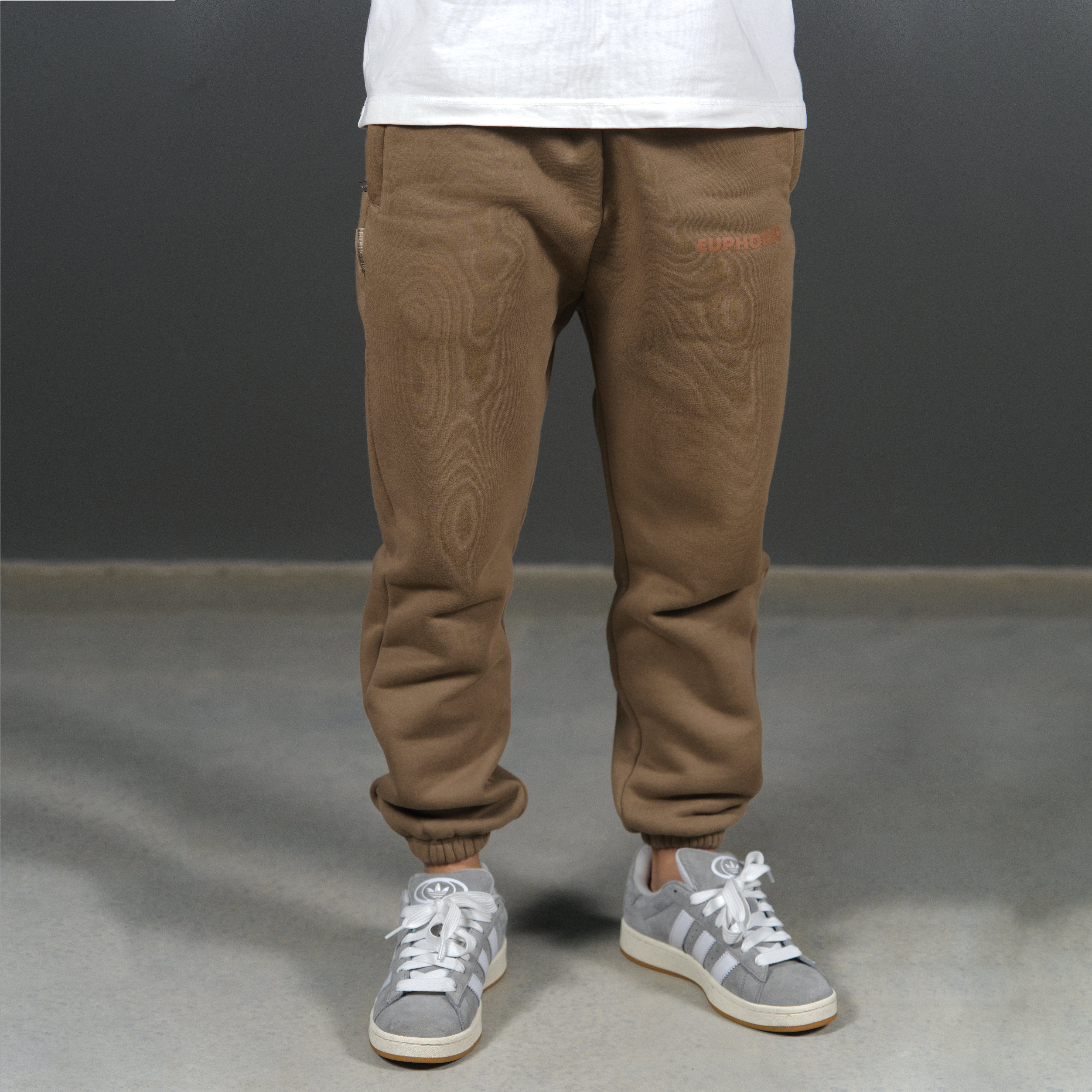 A person from the waist down wearing mocha sweatpants with elastic cuffs and grey sneakers, standing against a dark grey studio background.