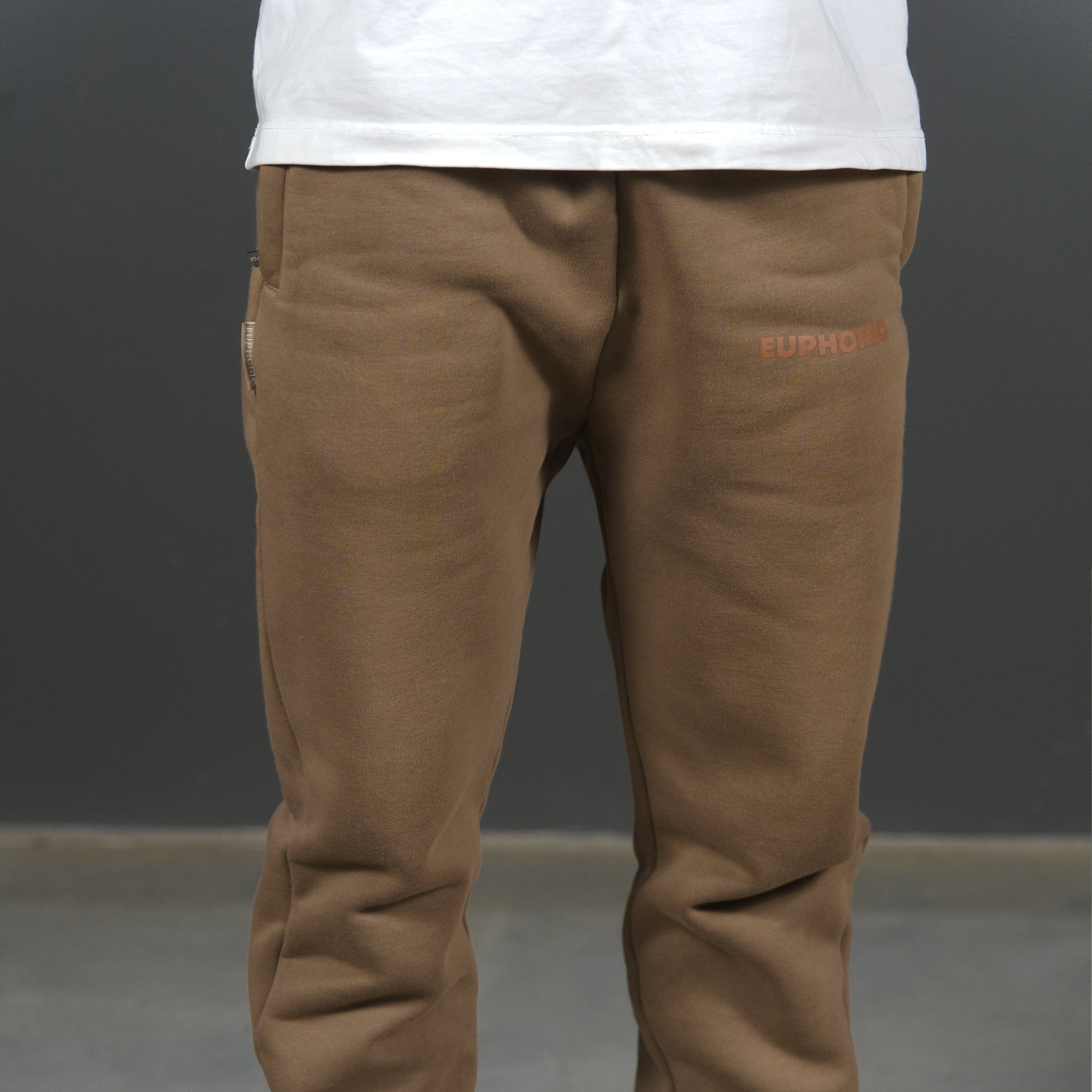 A person wearing mocha sweatpants with a small brown 'EUPHORIC' logo on the upper right thigh, paired with a white t-shirt, shown from the waist down against a dark grey studio background.