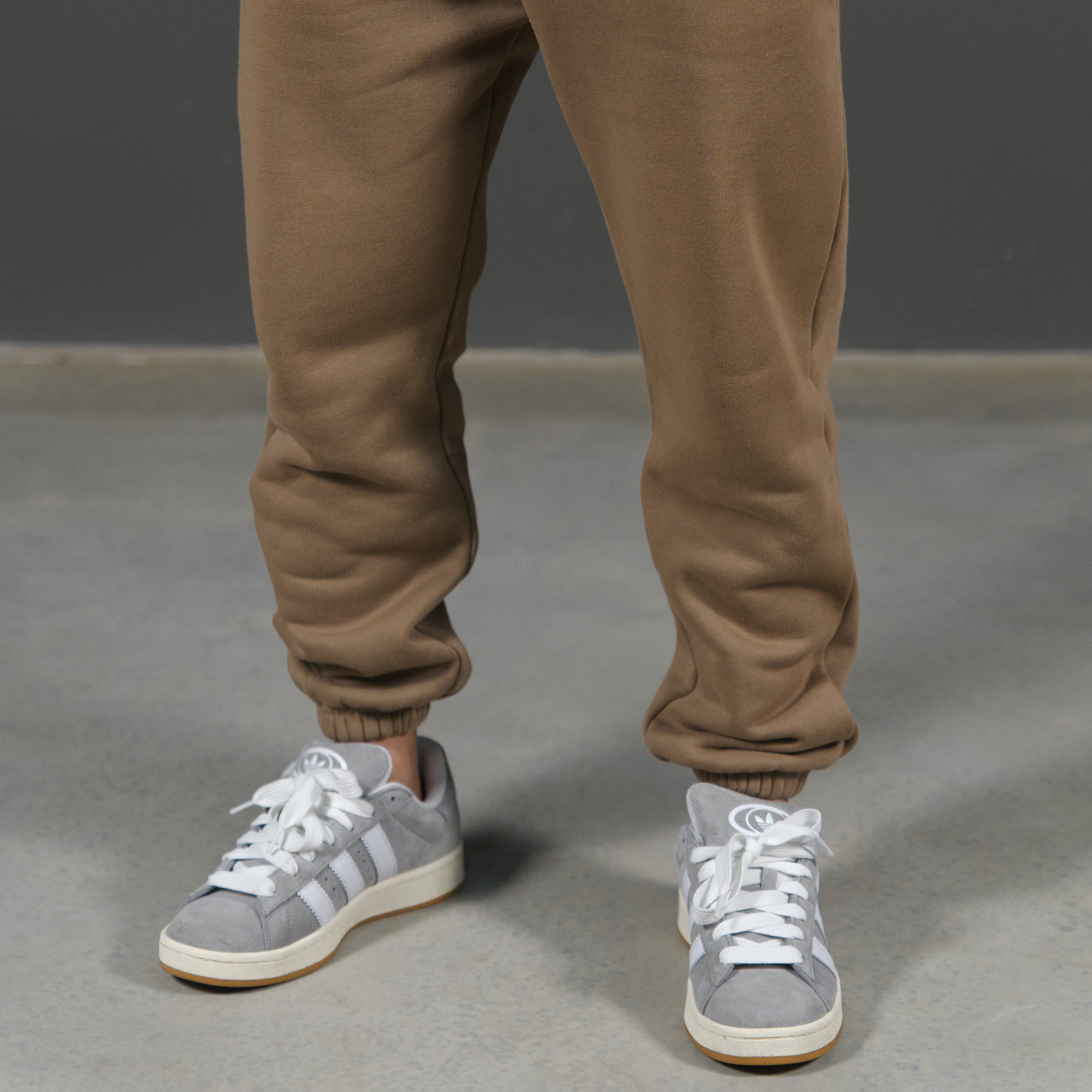 A person's legs wearing mocha sweatpants and grey low-top sneakers with white stripes, standing on a grey concrete floor against a dark grey background.