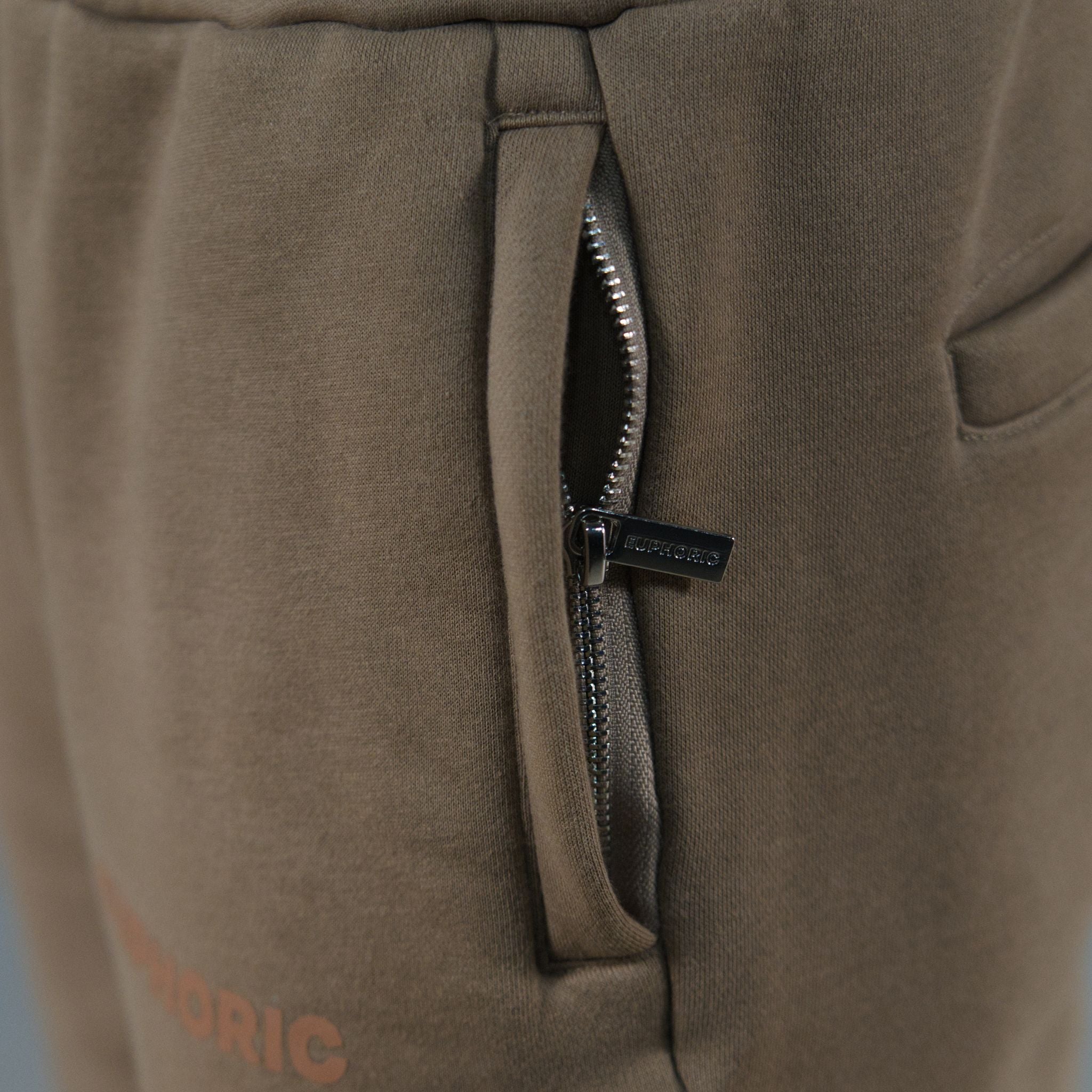 Close-up of a pair of mocha sweatpants showing a zippered pocket with a chrome zipper pull tab and partial brown text near the bottom left.