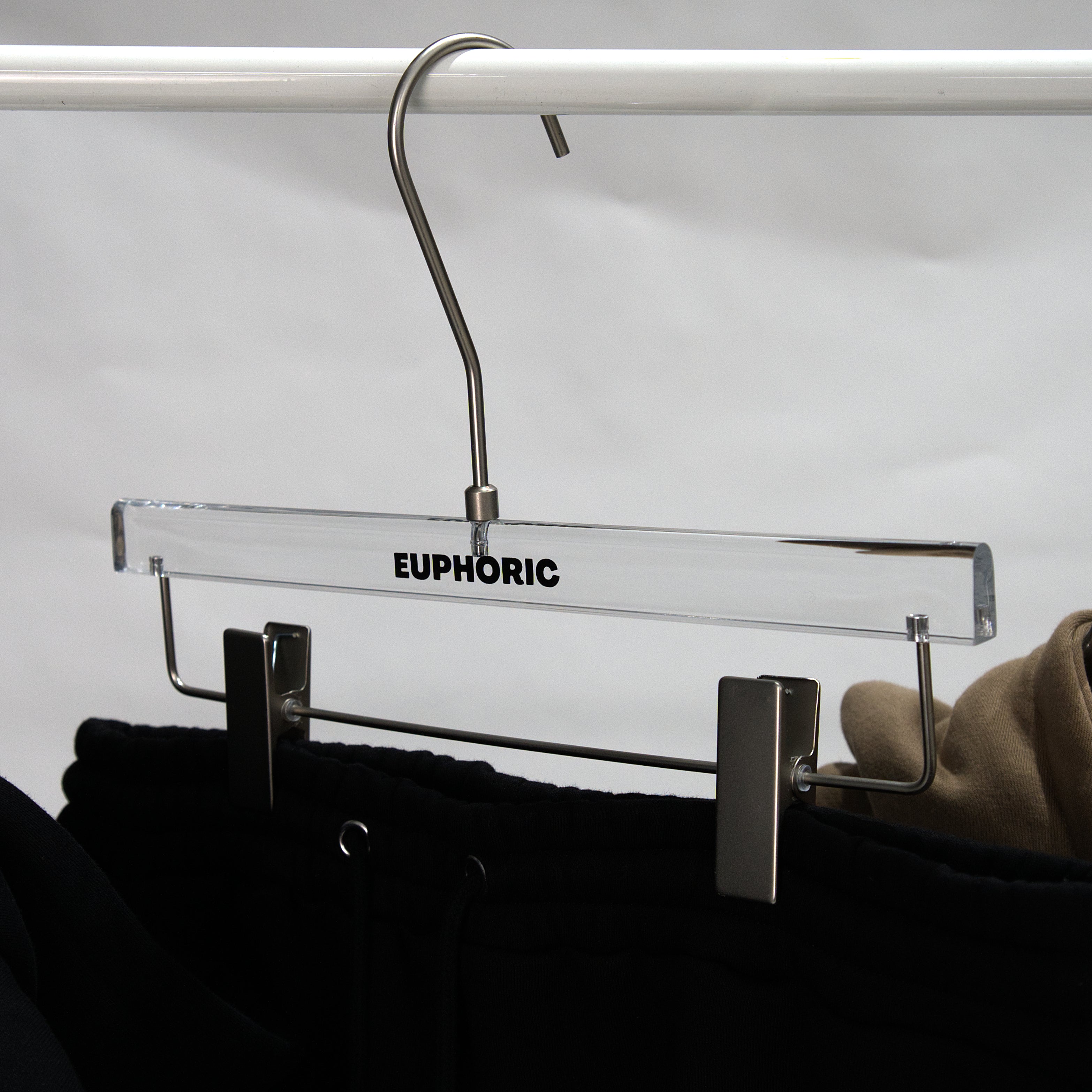 A clear acrylic clothes hanger with a silver metal hook and clips, featuring the brand name 'EUPHORIC' printed in black on the bar, holding black pants.